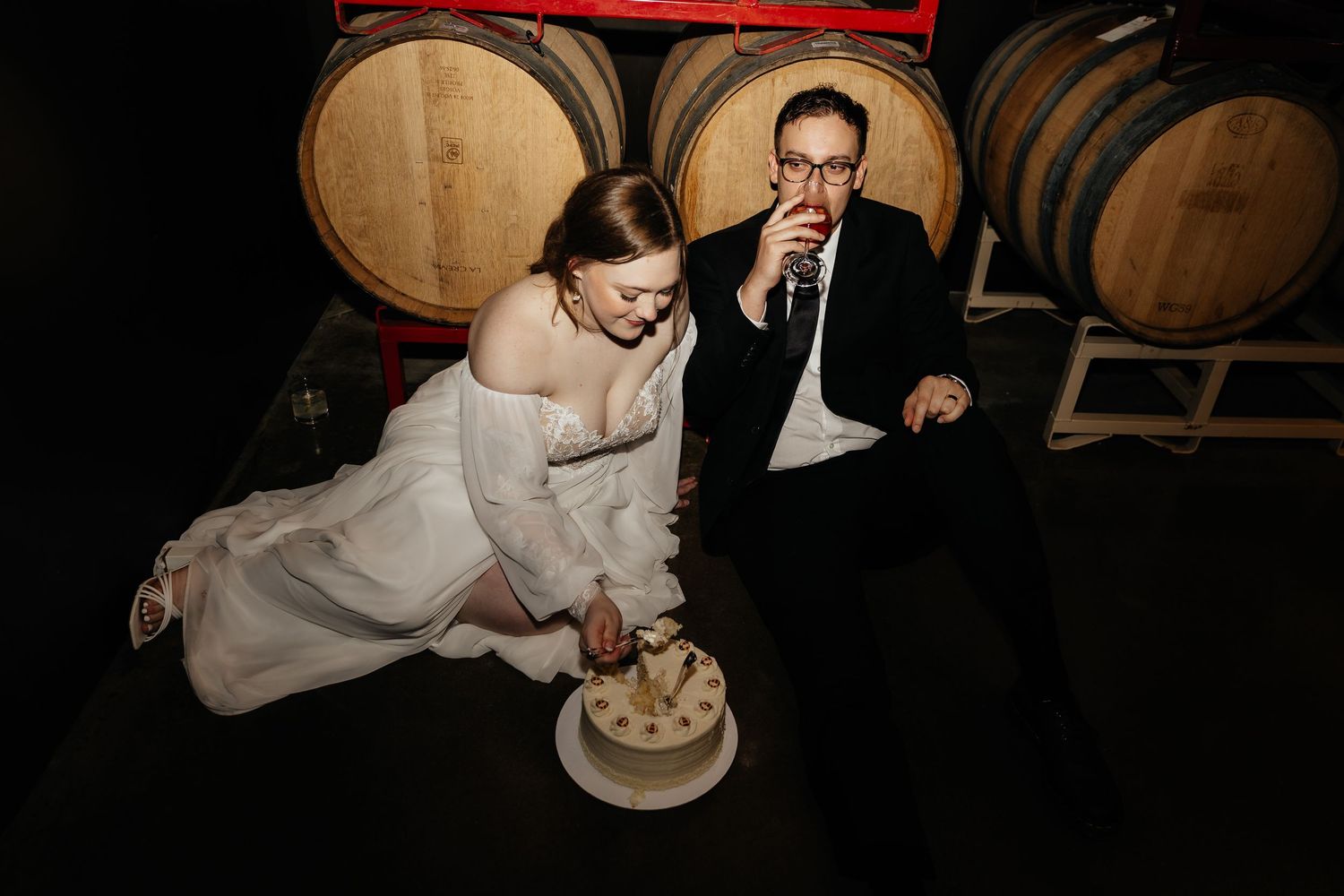 Bride and groom sit on floor cutting wedding cake beside wine barrels in dark rustic winery venue.