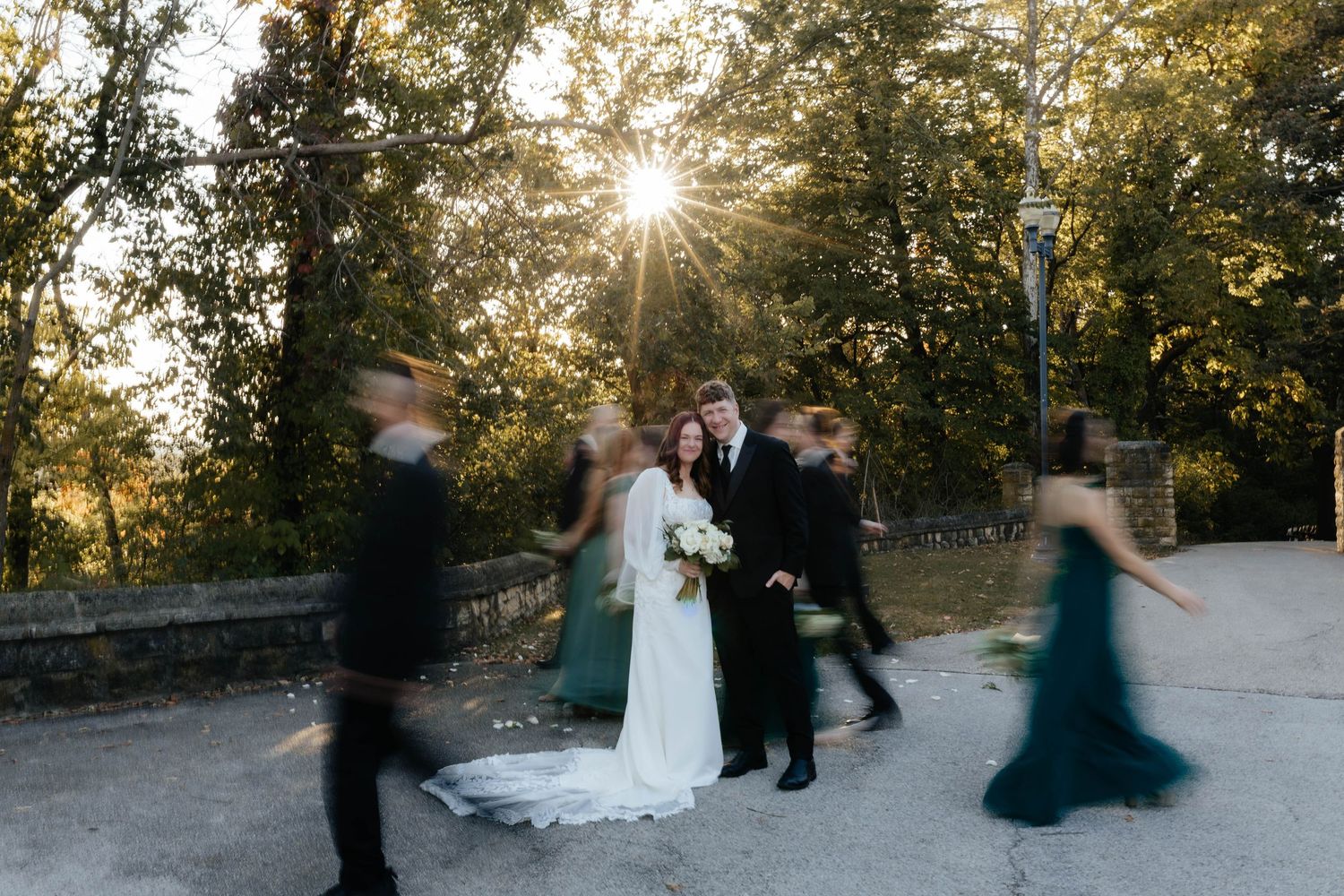 Bride and groom pose with wedding party at golden hour, sun rays bursting through trees as bridesmaids in green dresses blur past.