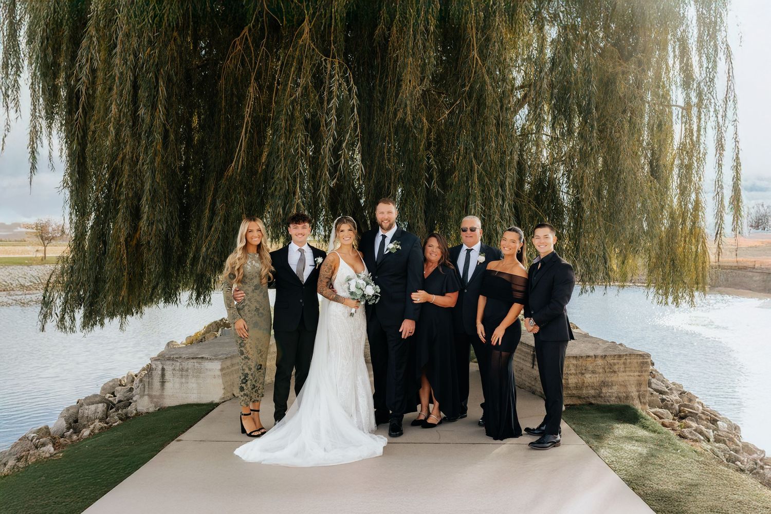 Wedding party group portrait with bride and groom surrounded by family under weeping willow tree.