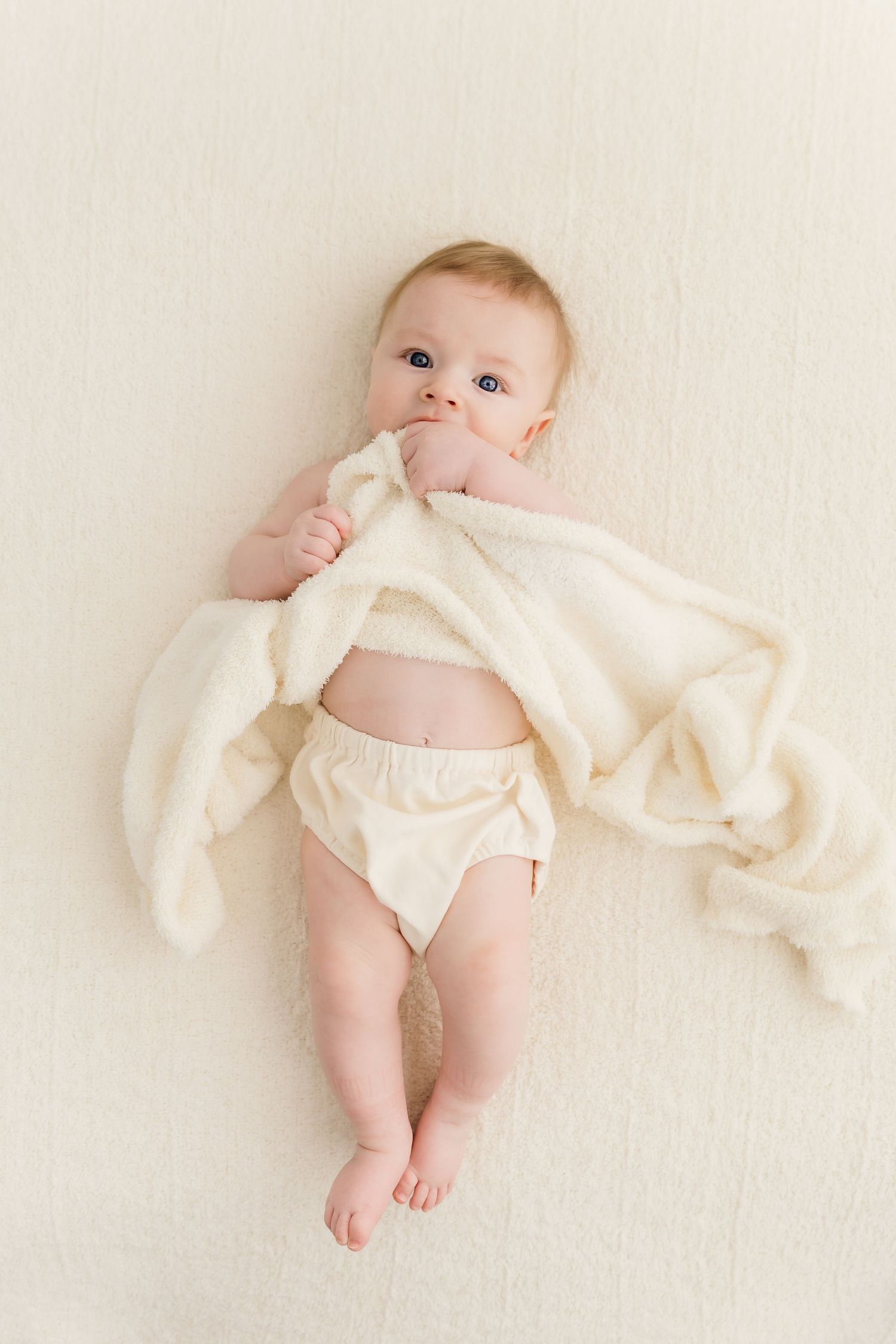 Sweet baby in diaper lying on back holding a cream fluffy blanket.