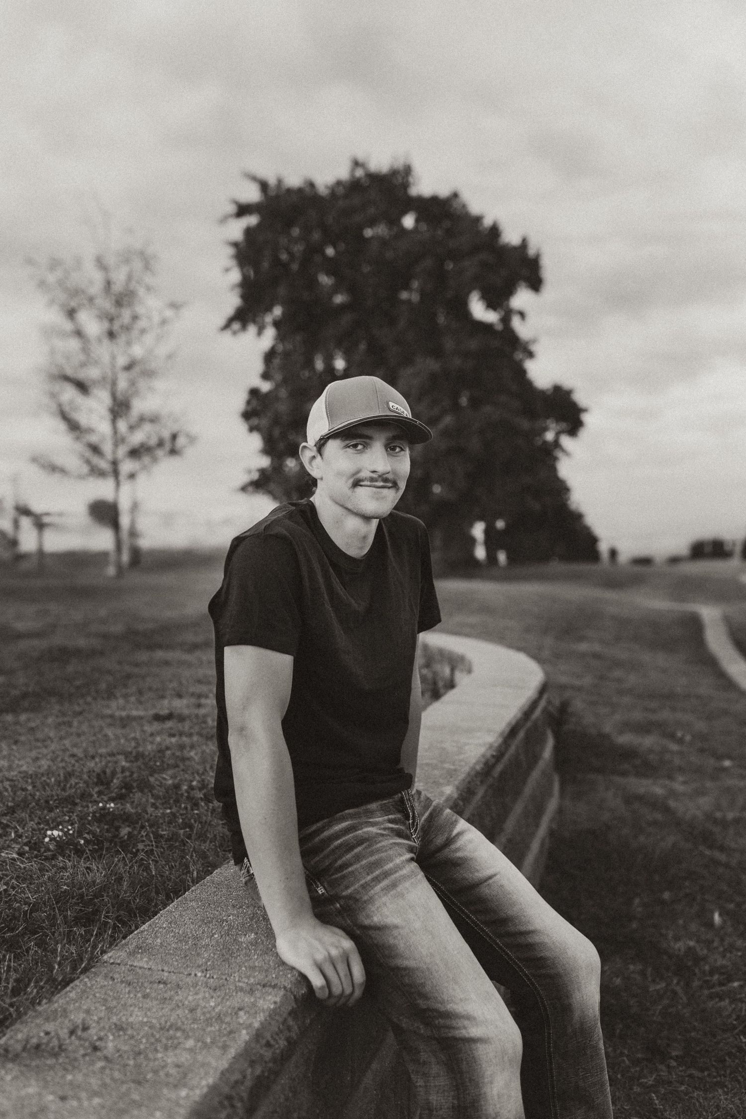 Black and white portrait of young man in cap and jeans sitting on a low wall outdoors.