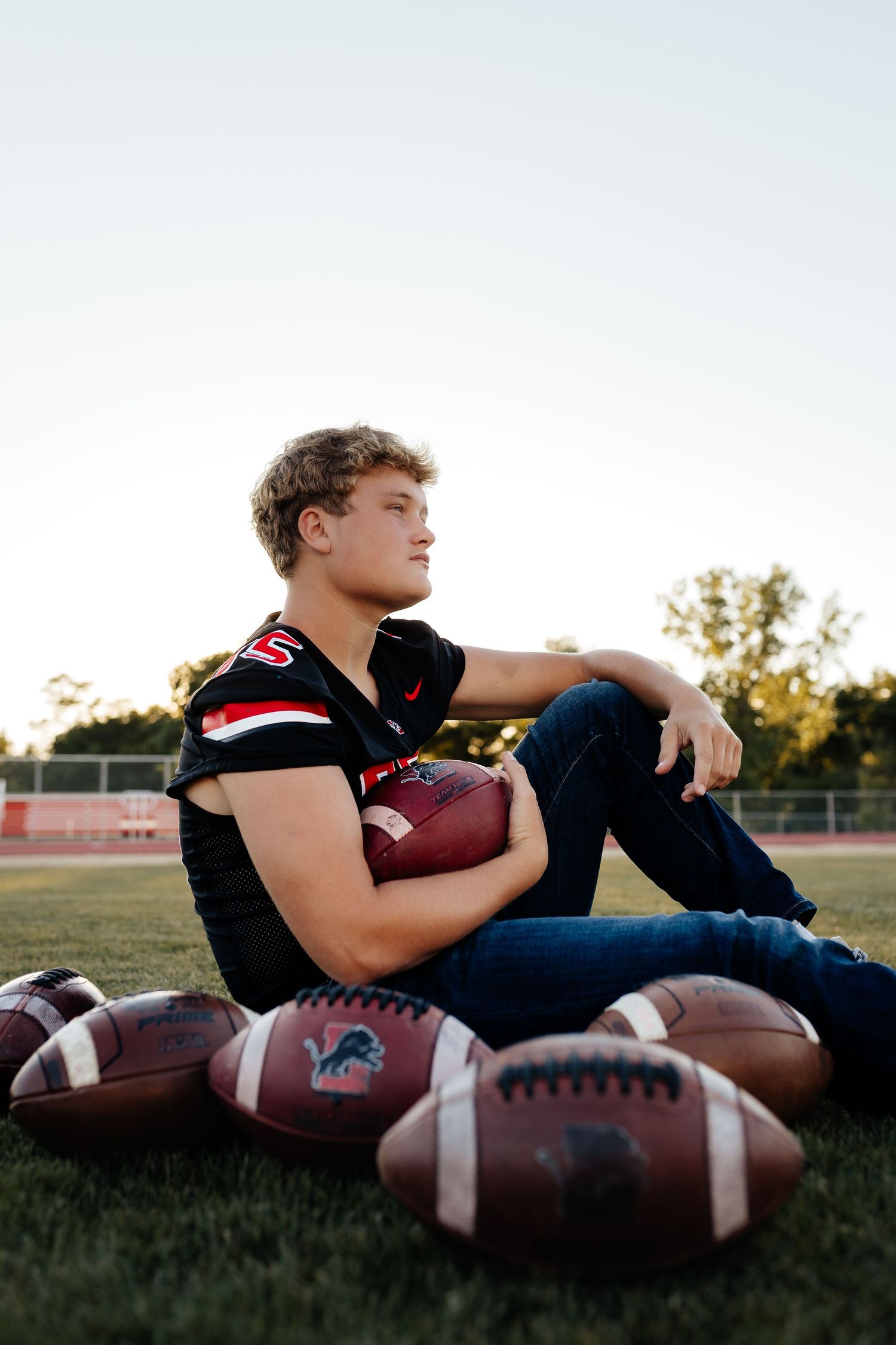 Teen athlete in football jersey sitting on grass surrounded by footballs at sunset.