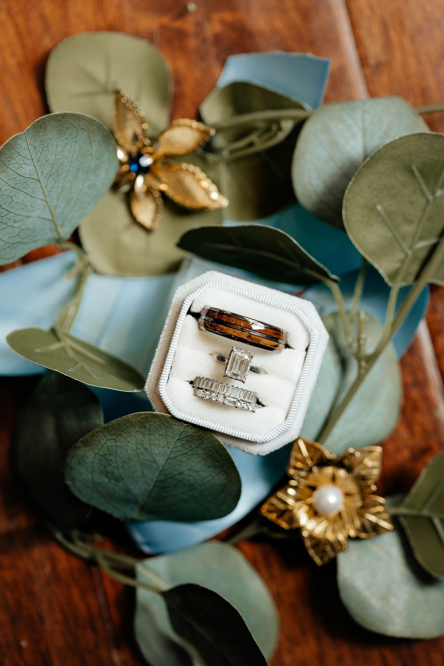 Wedding rings displayed in a white velvet box surrounded by eucalyptus leaves and gold floral accents.