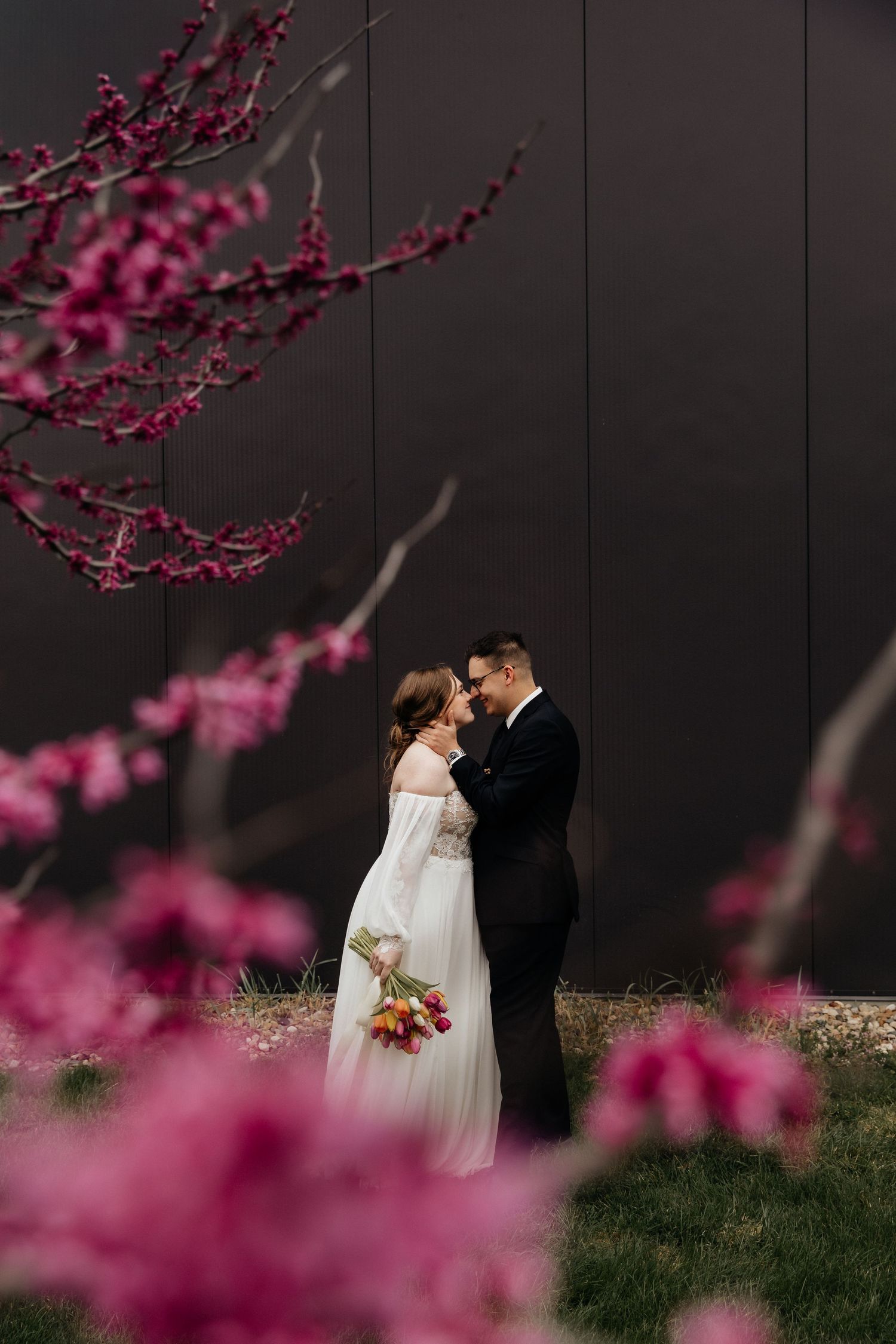 Bride and groom embrace tenderly surrounded by vibrant pink cherry blossoms against dark wall.
