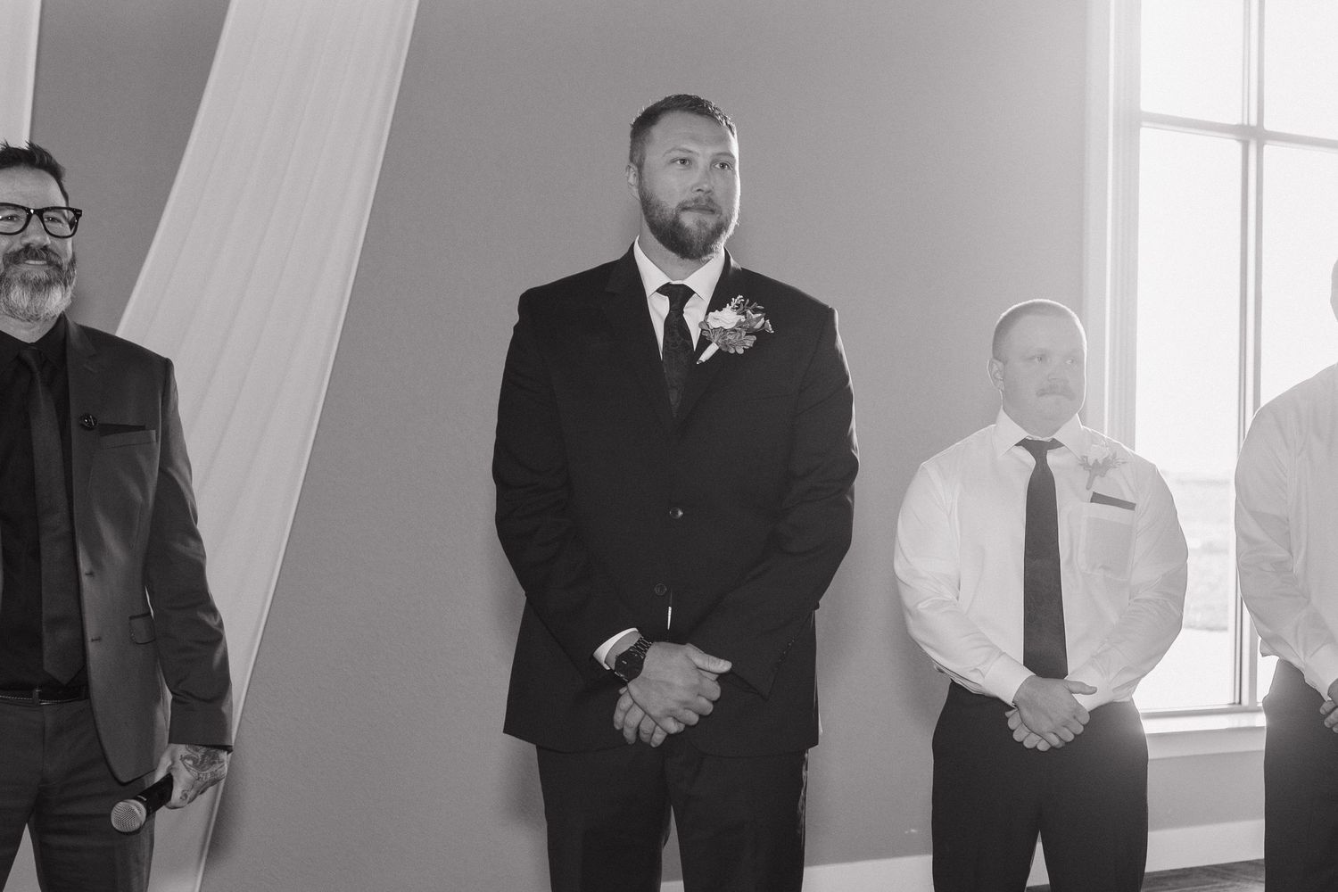 Groom in black suit with boutonniere stands waiting at wedding ceremony altar with guests nearby.