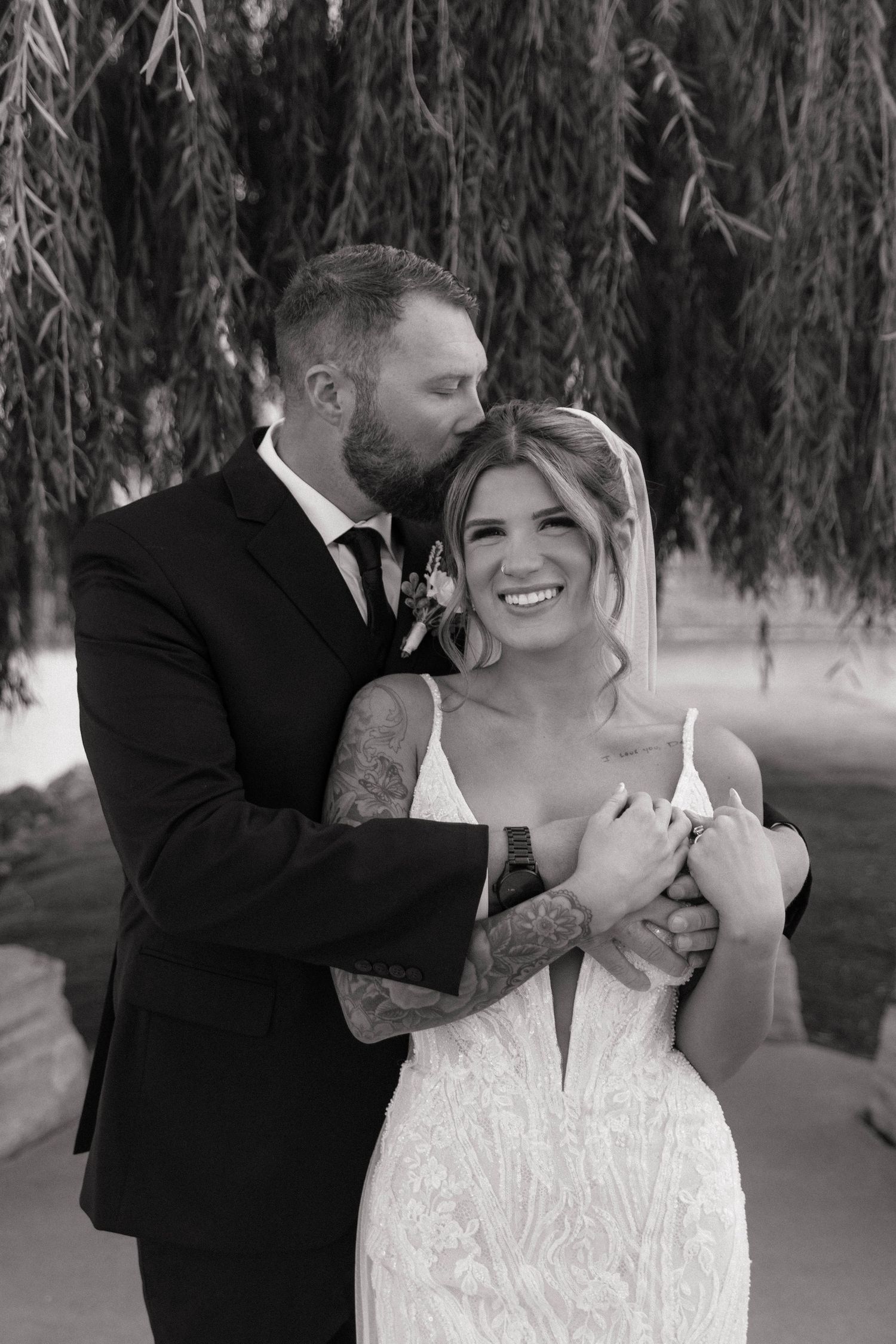 Black and white photo of a groom kissing his smiling bride on the cheek under a willow tree.