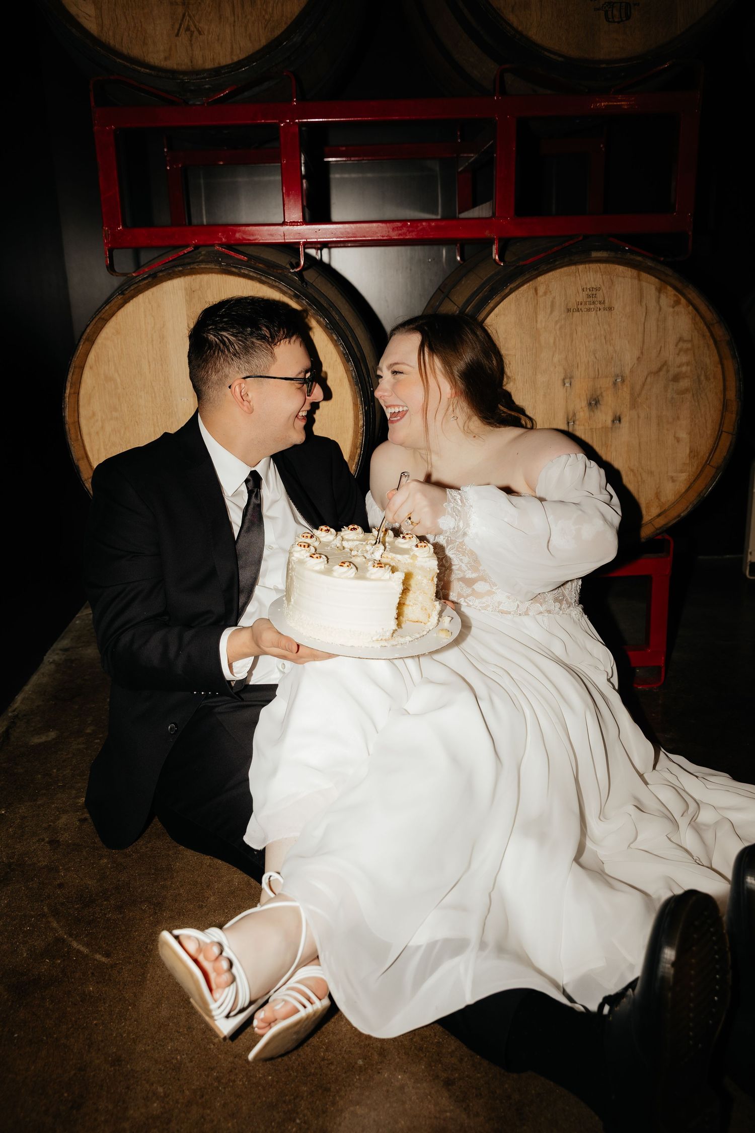 Smiling bride and groom sit together holding white wedding cake in front of wooden barrels at winery venue.