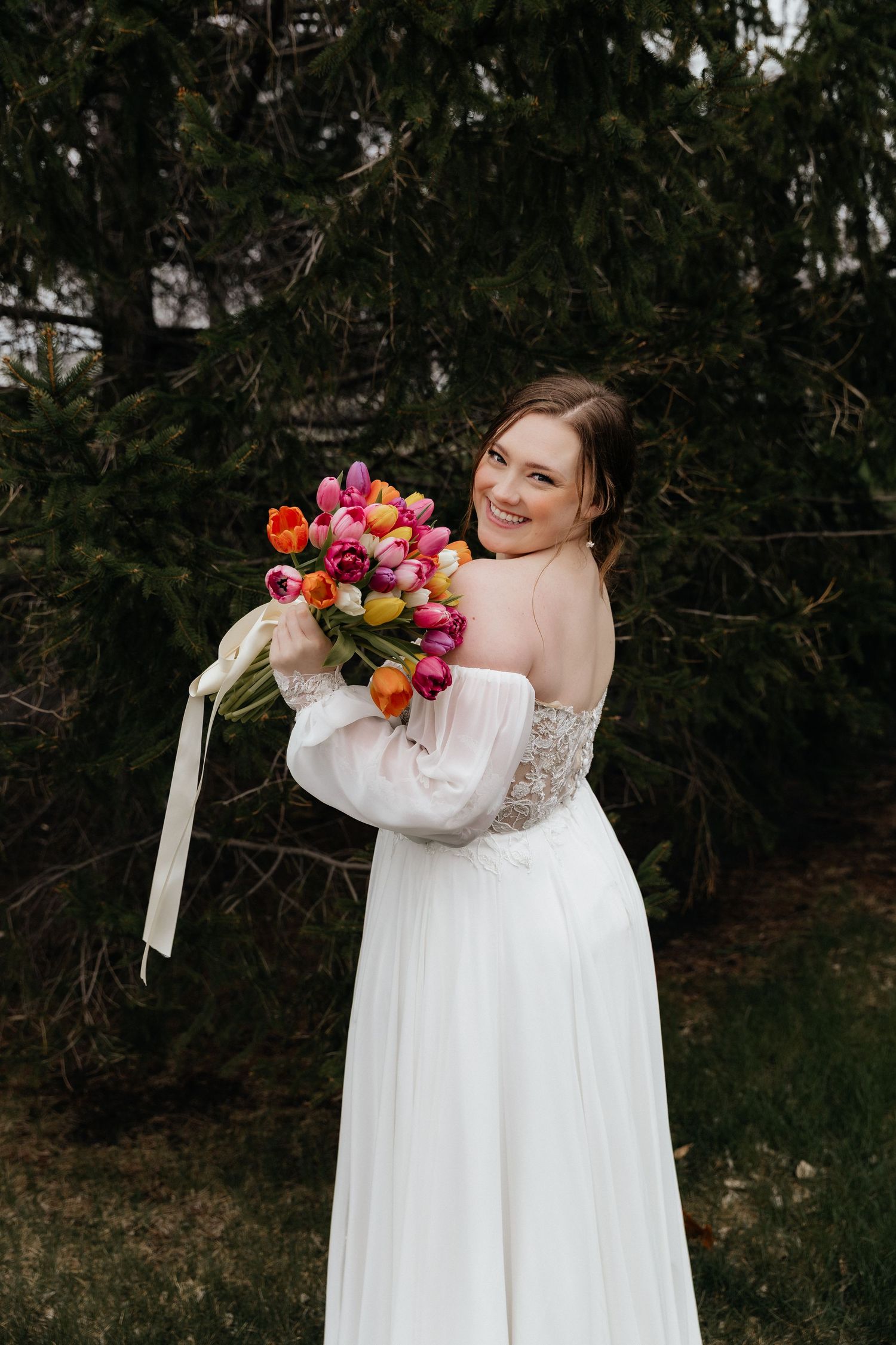 Joyful bride in off-shoulder lace gown holding colorful tulip bouquet while posing outdoors near evergreen trees.