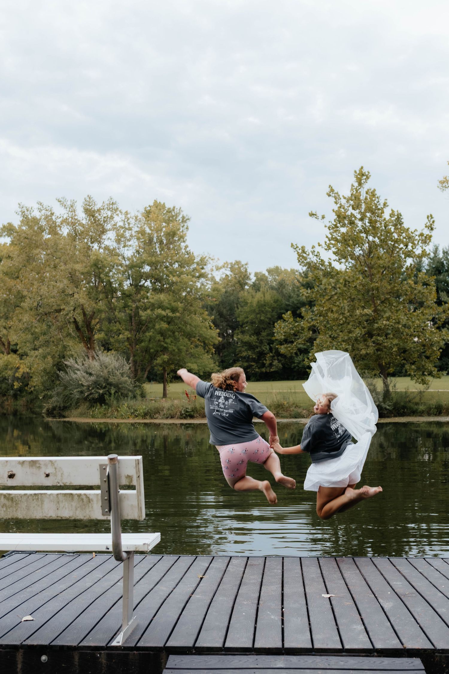 Bride in wedding dress and guest jumping off a dock into a lake holding hands.