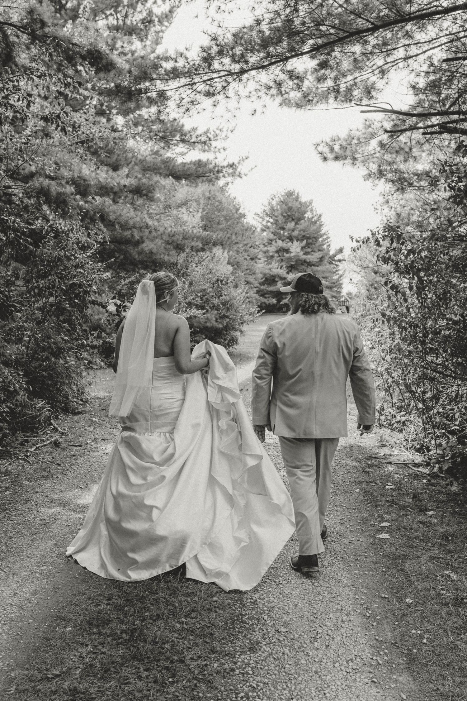 Black and white photo of newlyweds walking hand-in-hand down a tree-lined path after their ceremony.