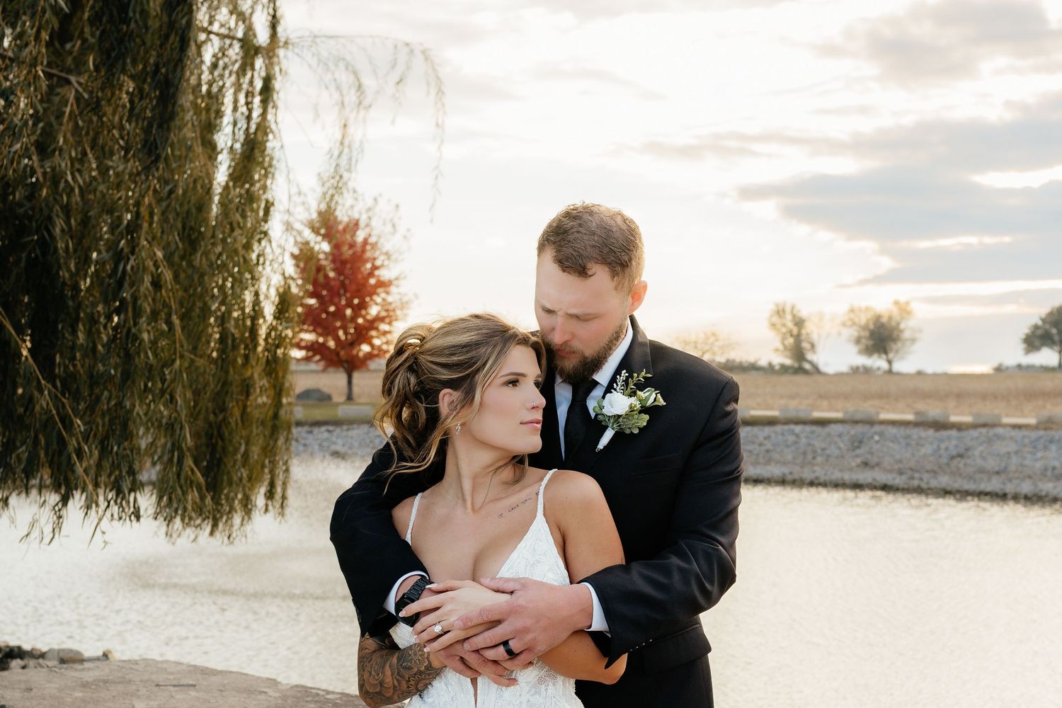 Bride and groom embrace near a willow tree by a pond at golden hour during their wedding.