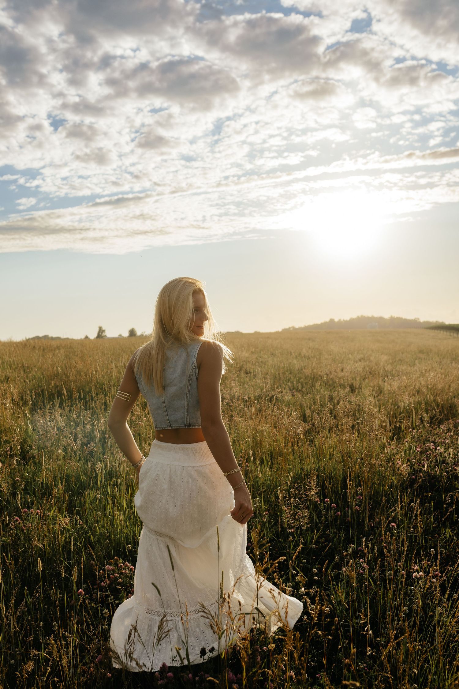 Blonde woman in white skirt and denim top standing in field at golden sunset.