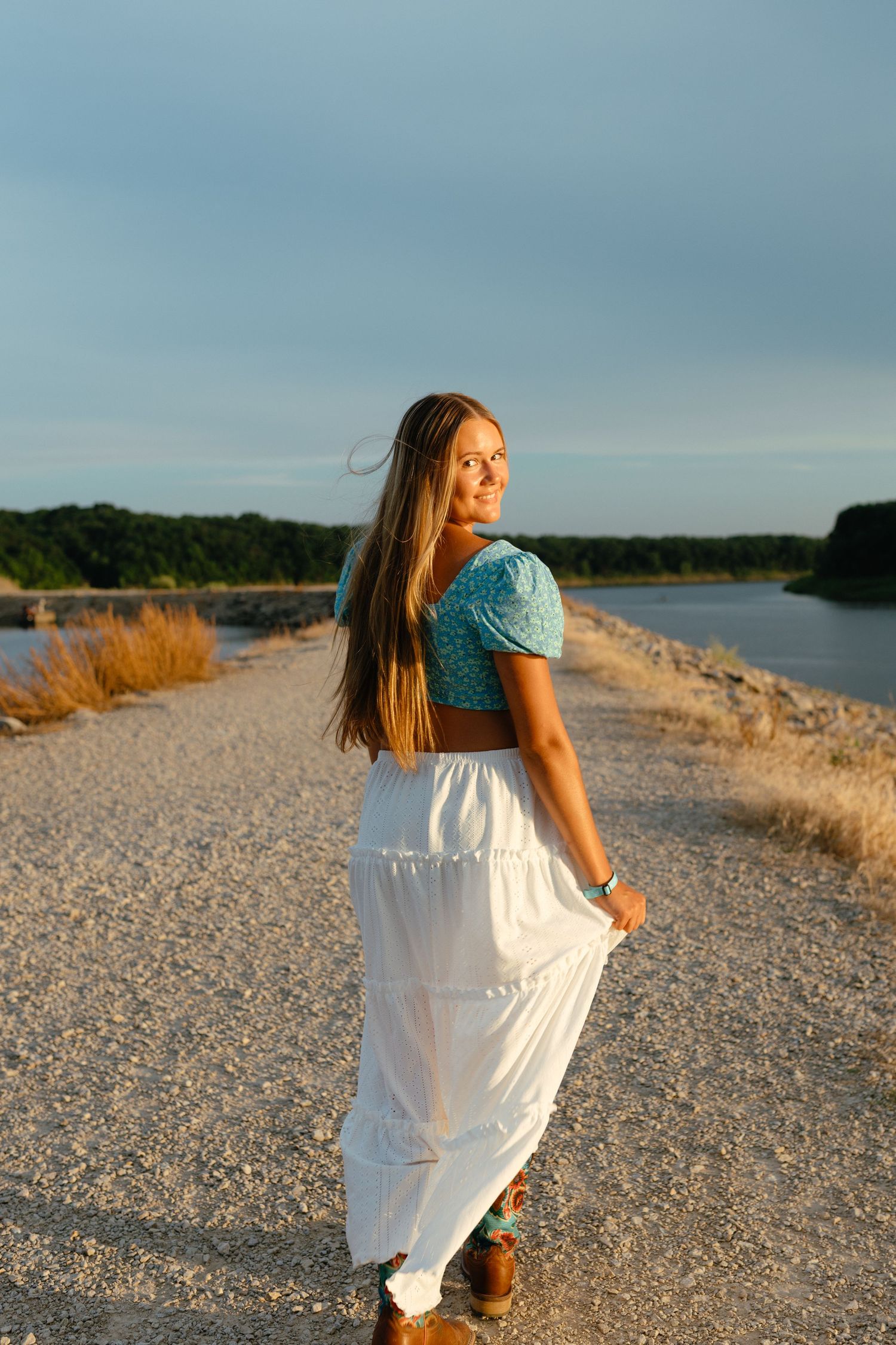 Blonde girl in blue top and white skirt walks on gravel path along scenic lake at sunset.