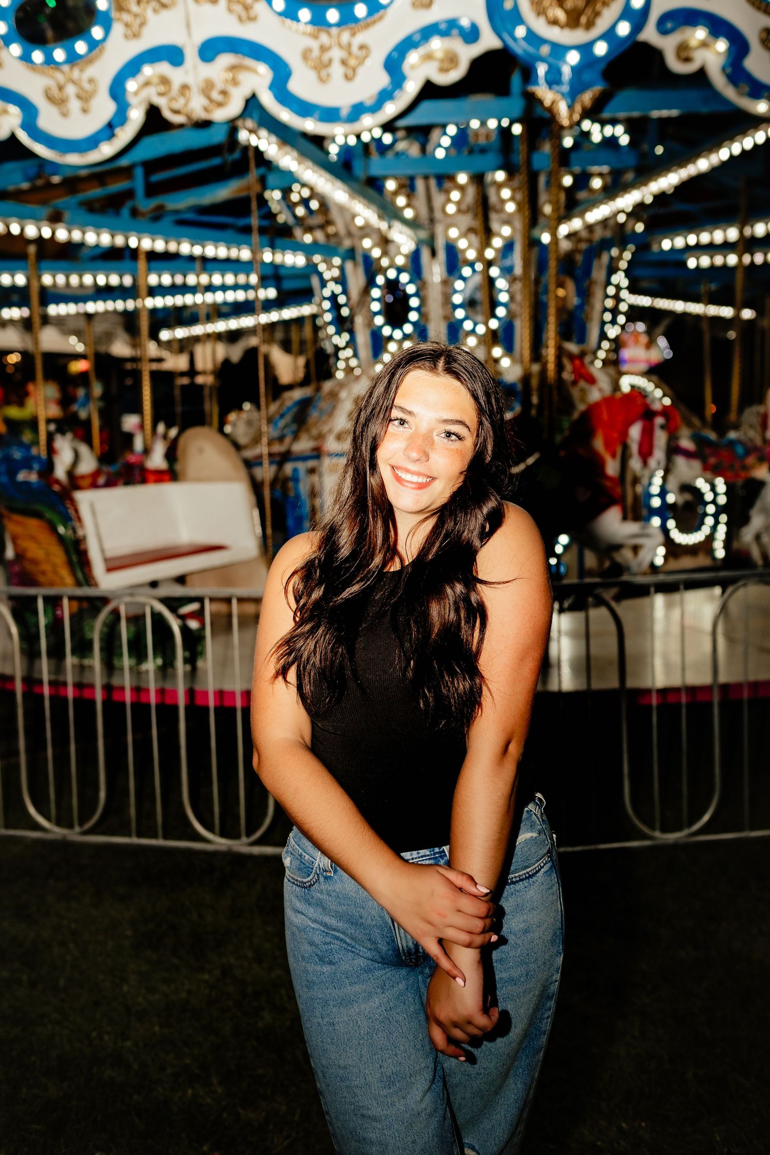Young woman in black top and jeans smiling in front of a lit carousel at night.
