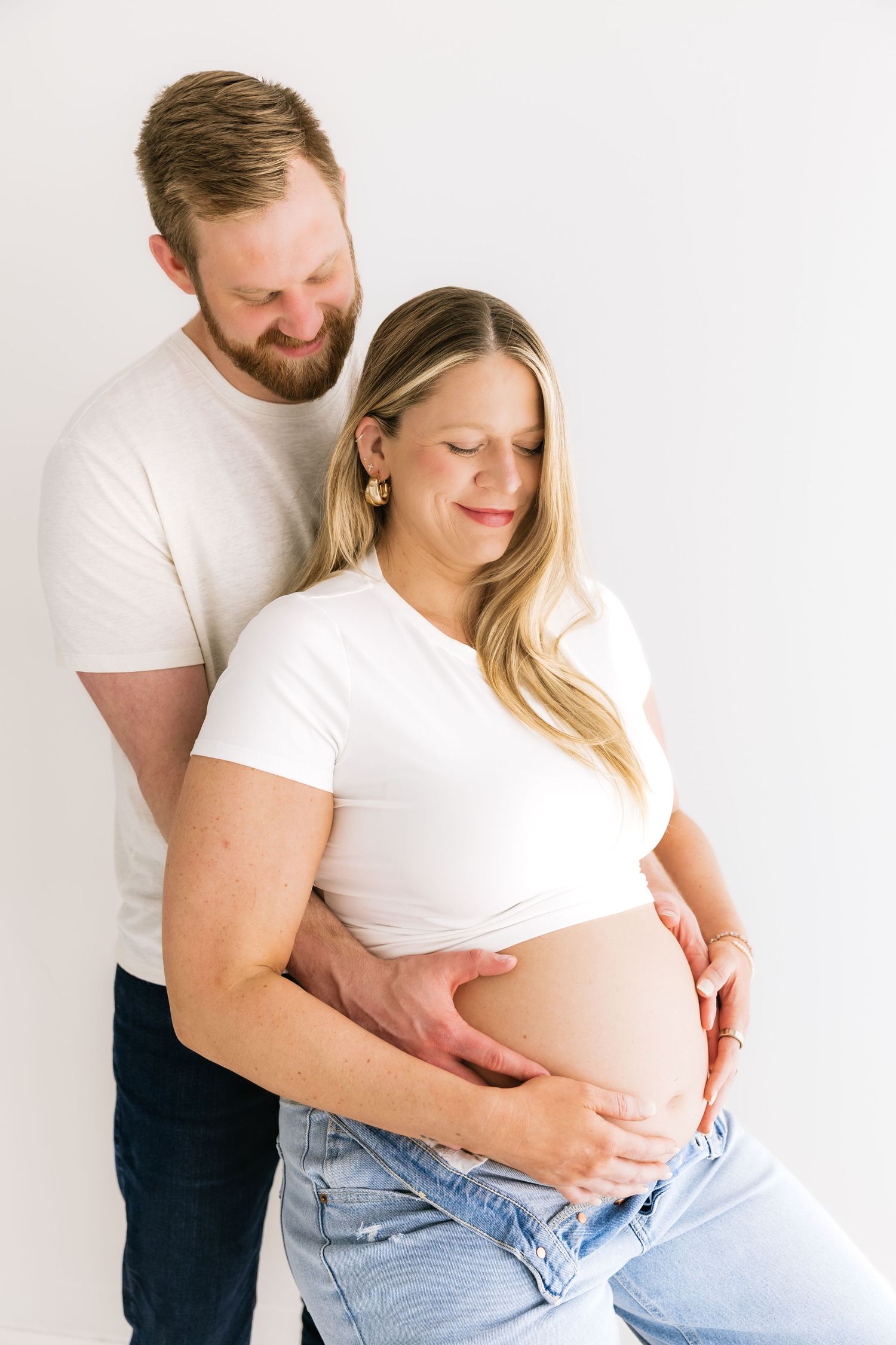 Expecting couple in white tops tenderly cradle pregnant belly in bright minimalist studio setting.