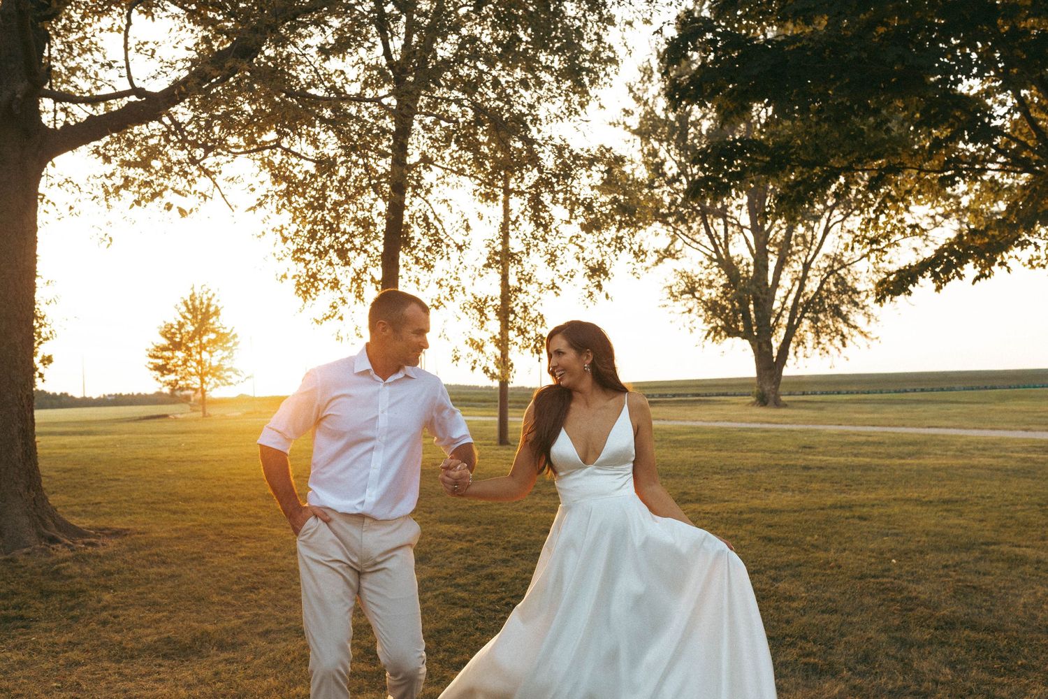 Couple holding hands walking through golden sunset light on a green open field.