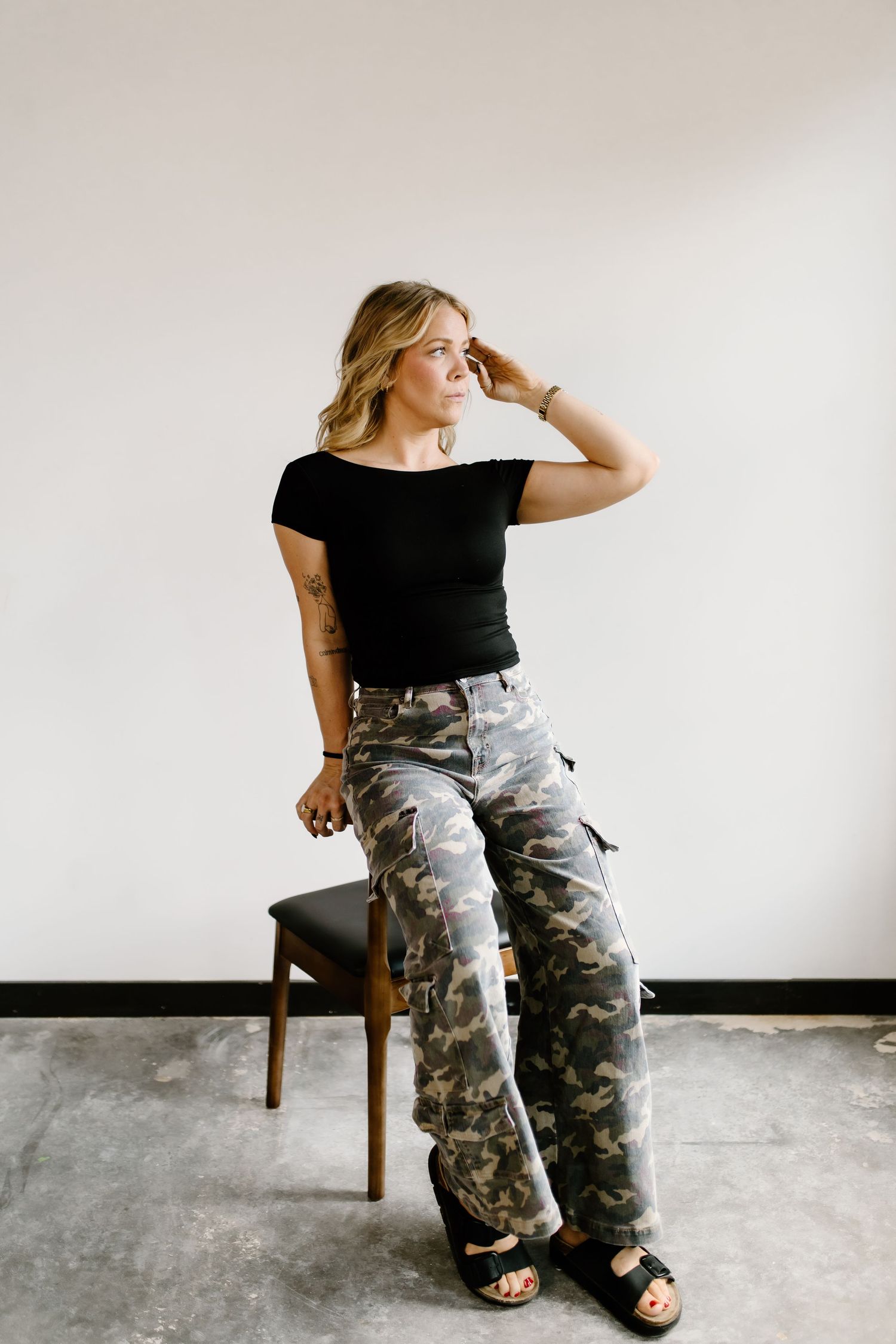 Confident blonde woman in black top and camo cargo pants posing on a chair in a studio.