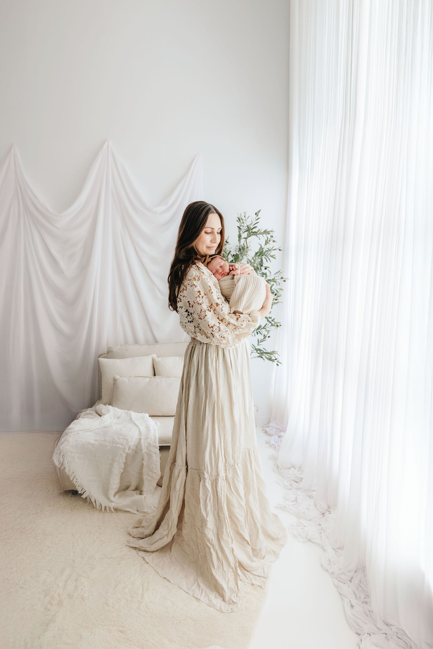 Mother in elegant cream lace gown tenderly holds newborn baby near bright window in white studio.