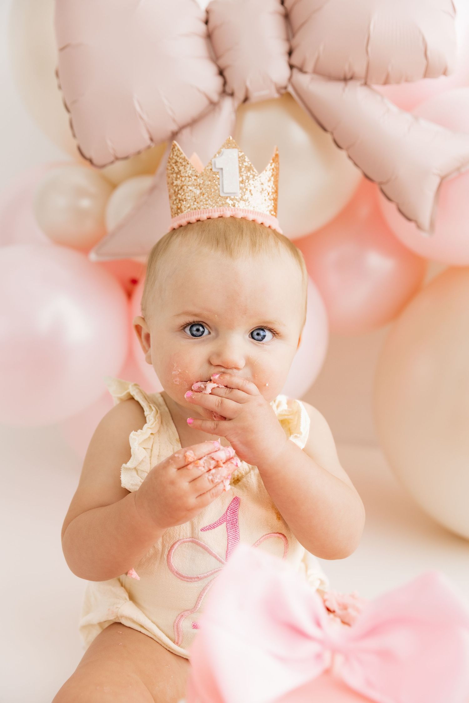 One year old baby girl in crown eats smash cake with pink balloons in background.