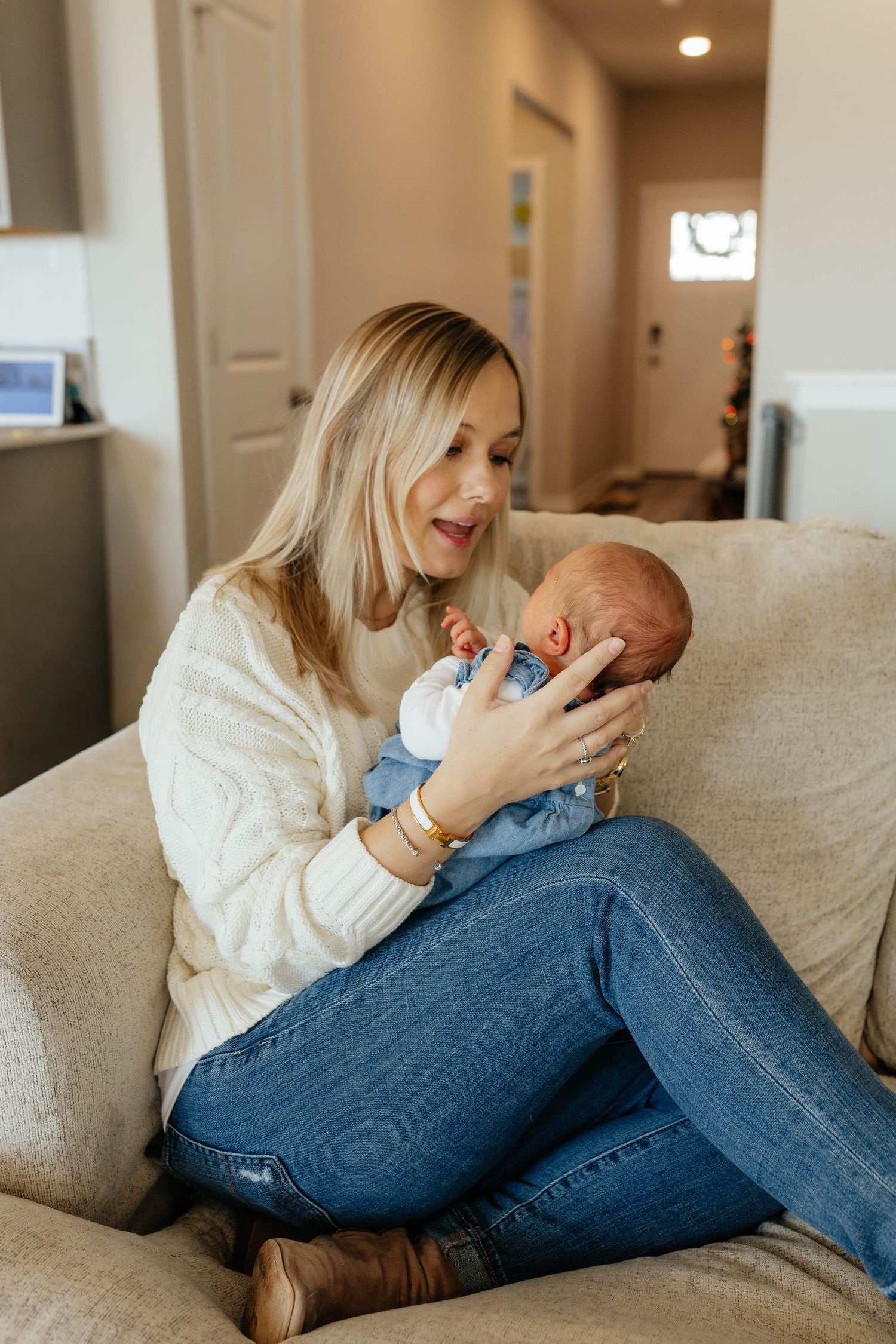A joyful blonde mother in a white sweater sits on a couch, gazing lovingly at her newborn baby.
