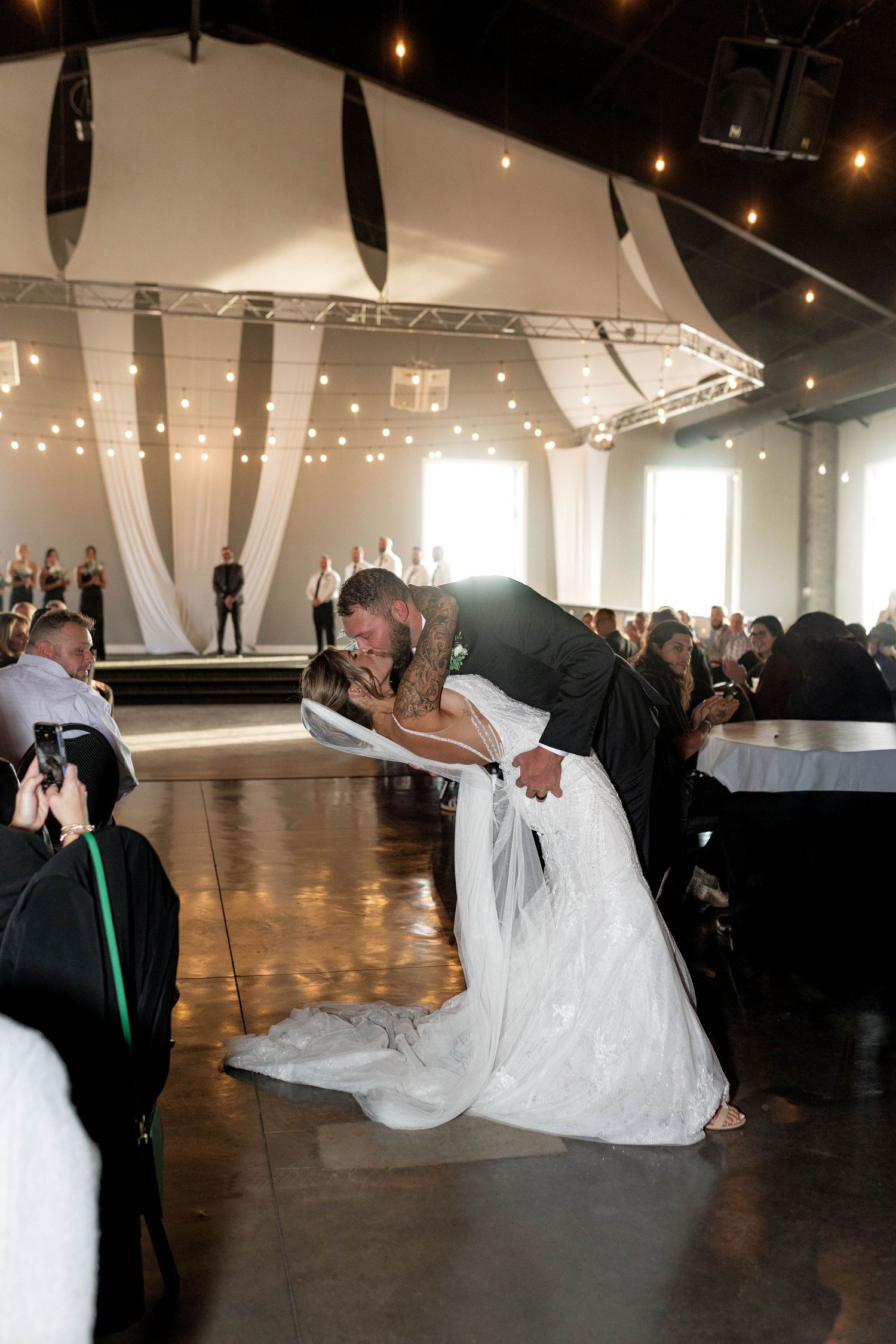 Groom dips bride for a romantic kiss on the dance floor during wedding reception with string lights.