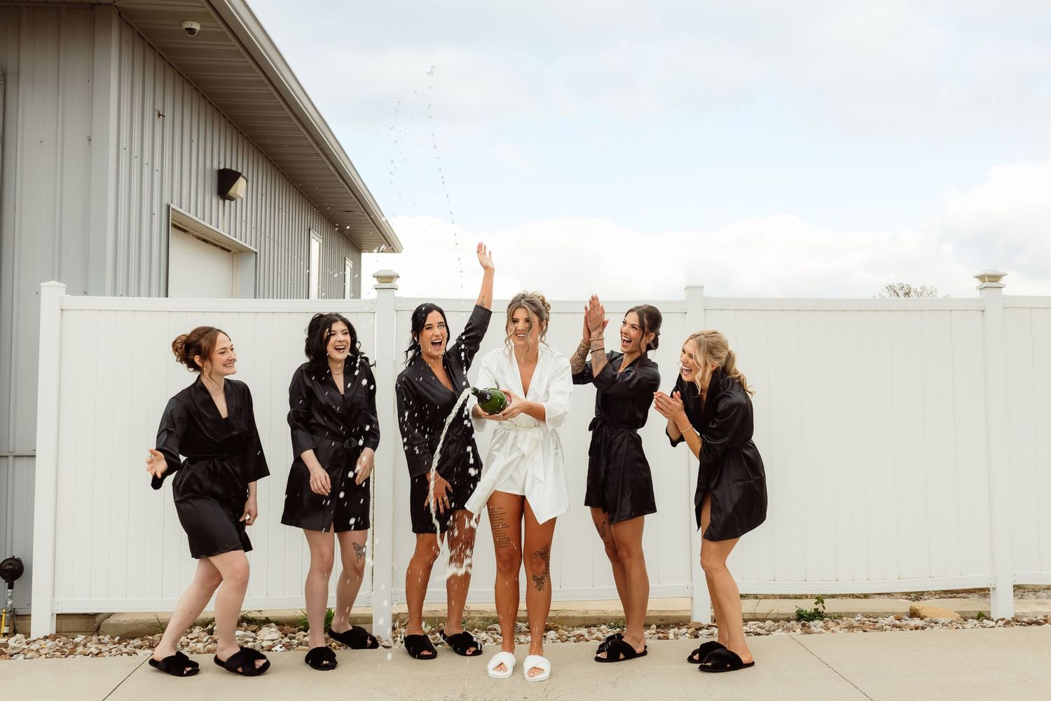 Bride celebrating with bridesmaids in black robes as champagne bubbles fly in the air.