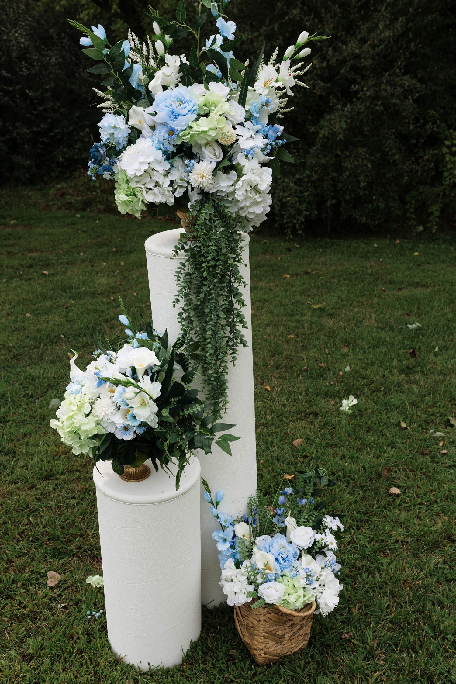 Blue and white floral arrangements on white pedestals and wicker basket for outdoor wedding ceremony.