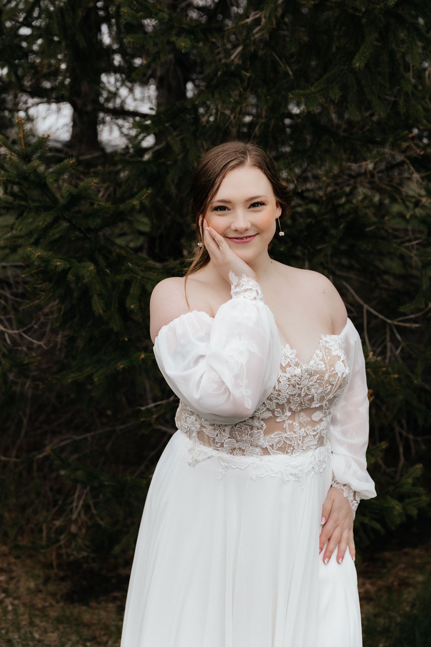 Elegant bride in off-shoulder lace wedding gown posing with hand on cheek smiling outdoors near evergreen trees.