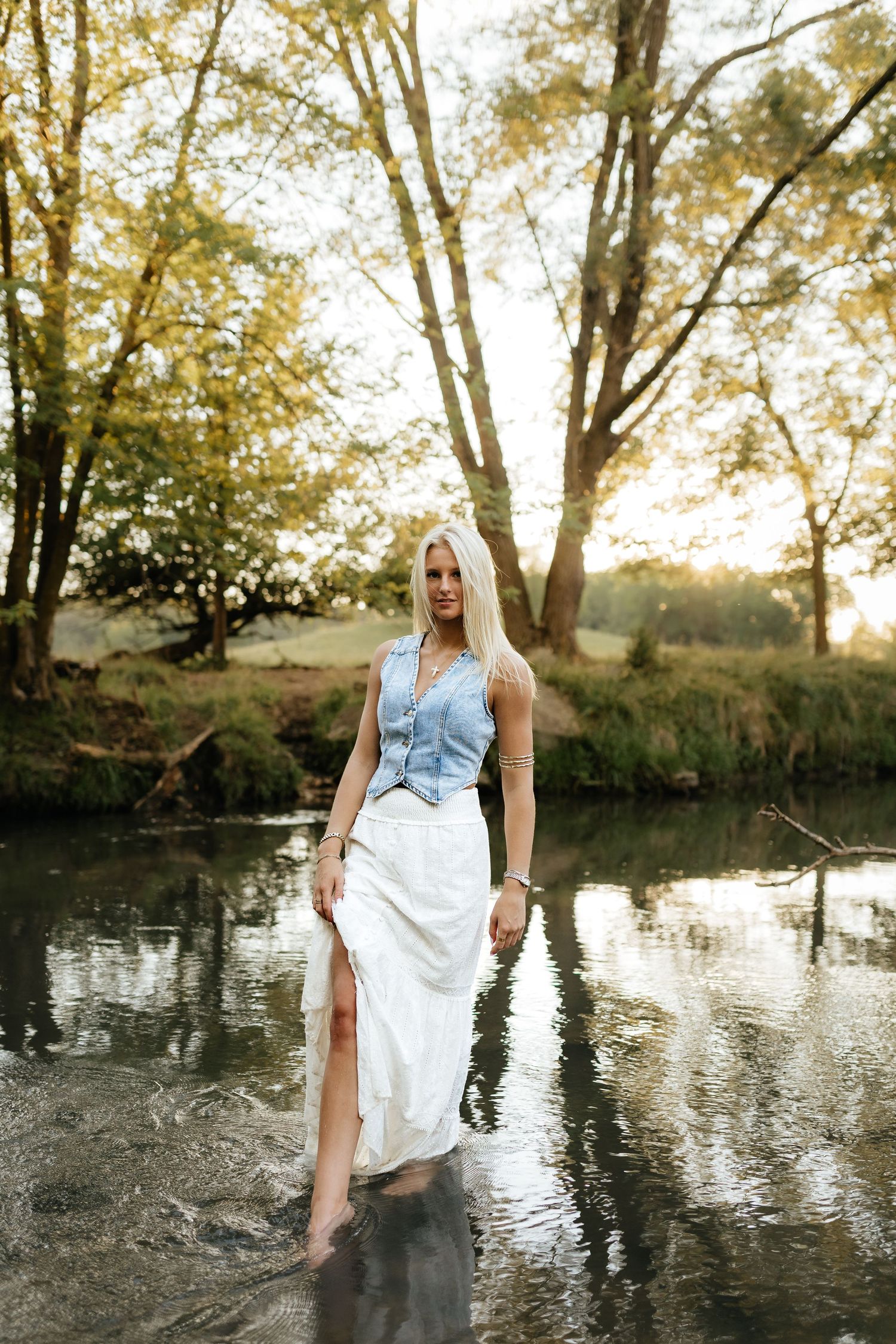 Blonde woman in denim vest and white skirt wading through calm creek in forest.
