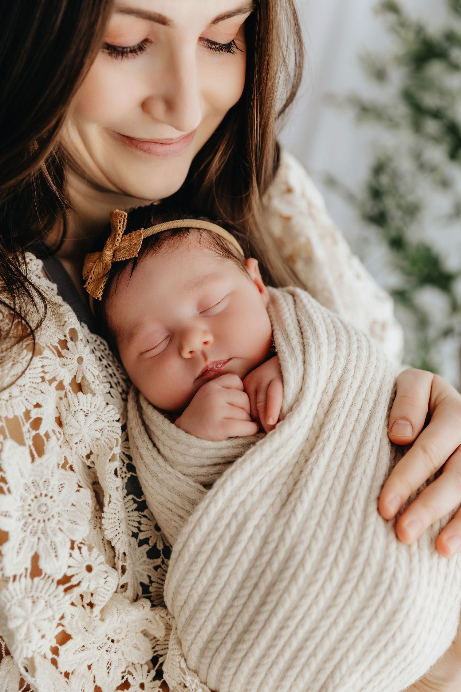 Mother in lace dress tenderly holds sleeping newborn wrapped in cream knit blanket with gold bow headband.
