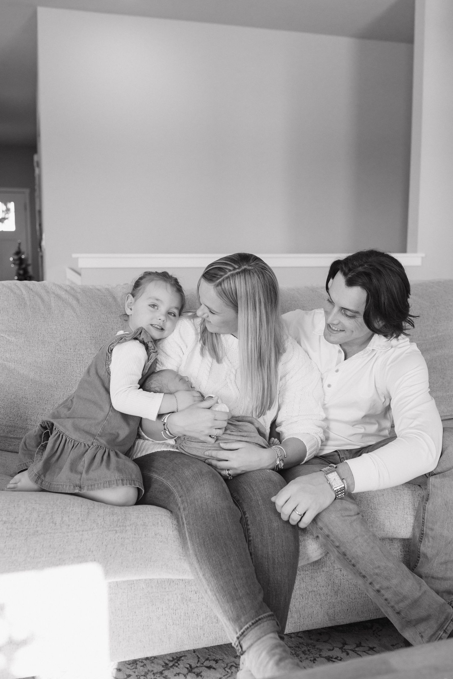 Black and white family portrait of parents and toddler daughter sitting on a couch with their newborn baby.