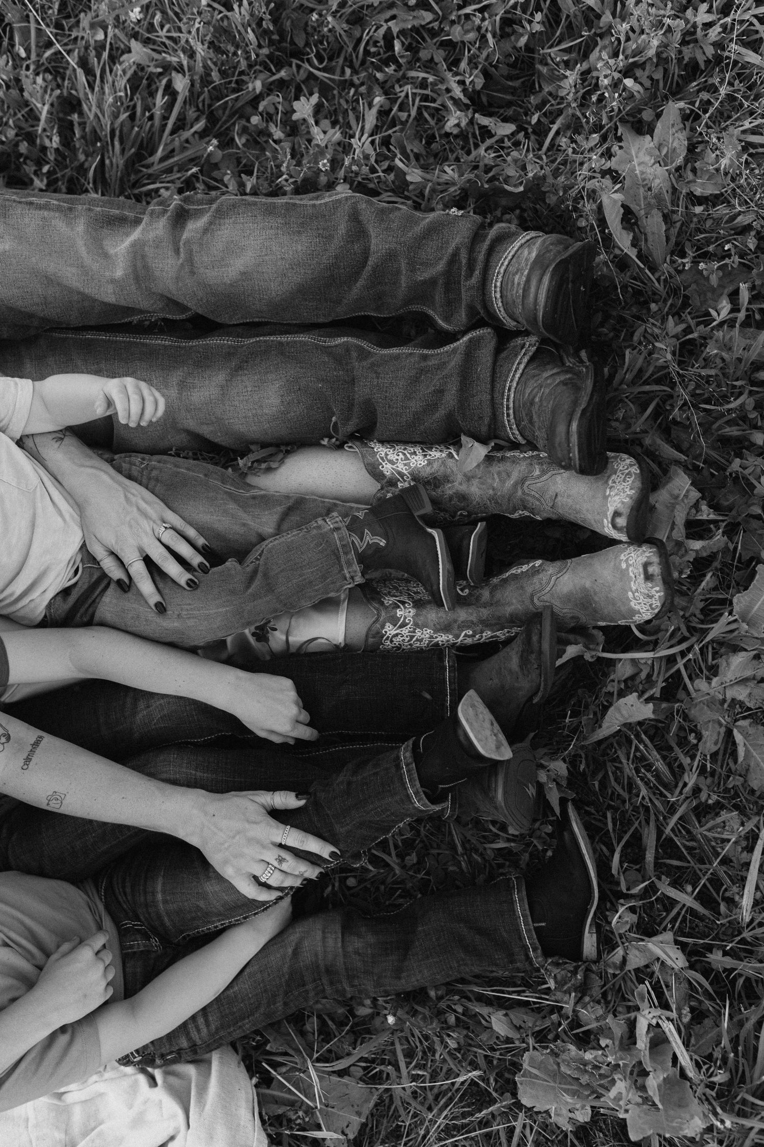 Black and white photo of family lying in grass showing cowboy boots and jeans with hands touching.