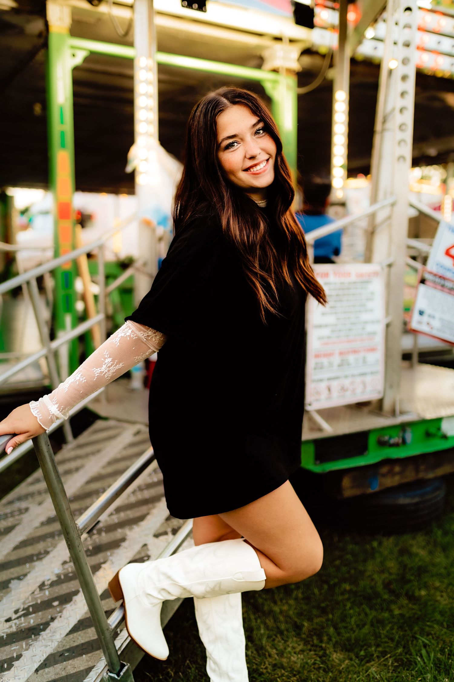 Young woman in black dress and white boots posing on steps of a colorful carnival ride.