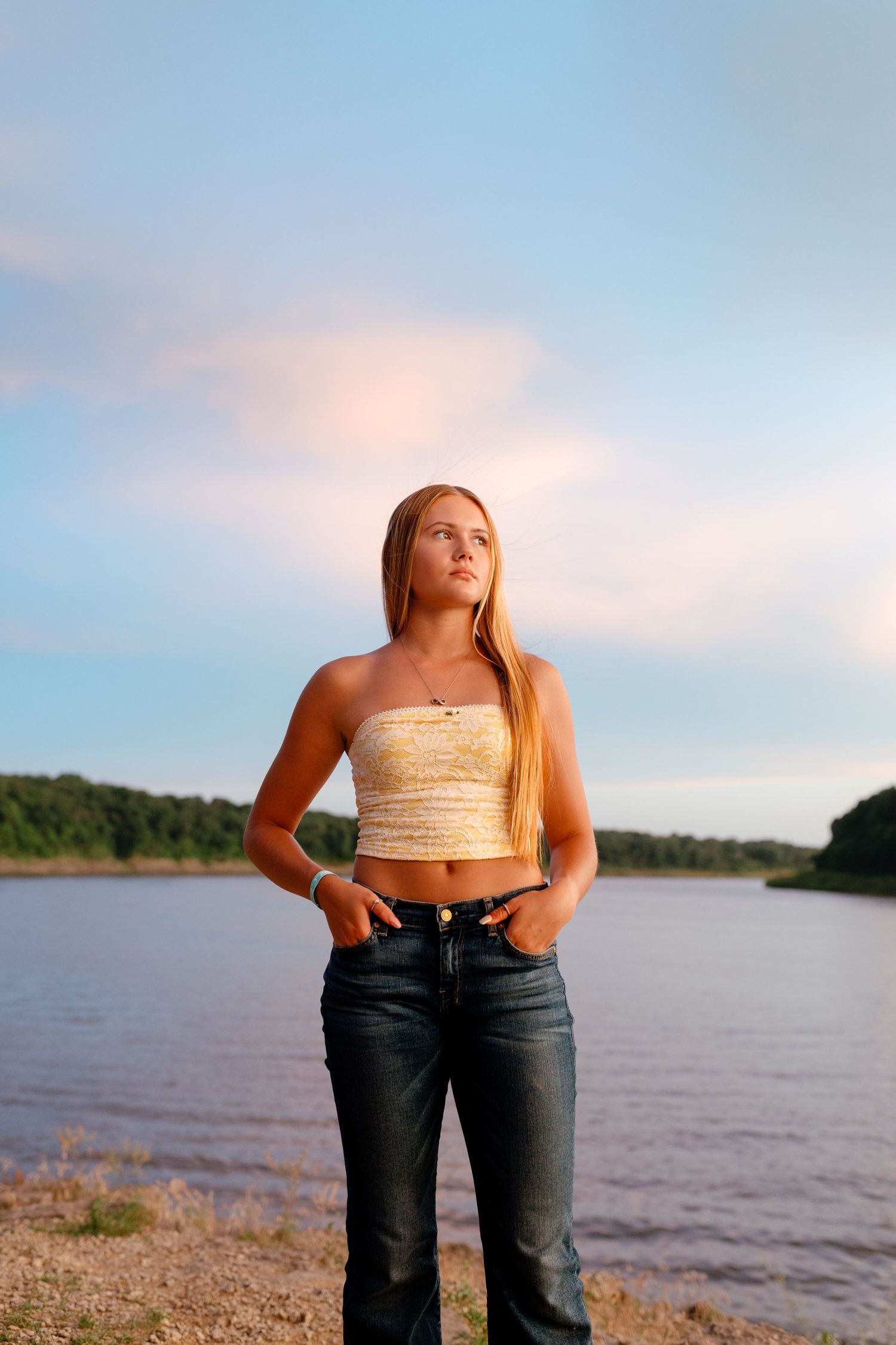 Young blonde woman in tube top and jeans poses by a river at sunset with hands in pockets.