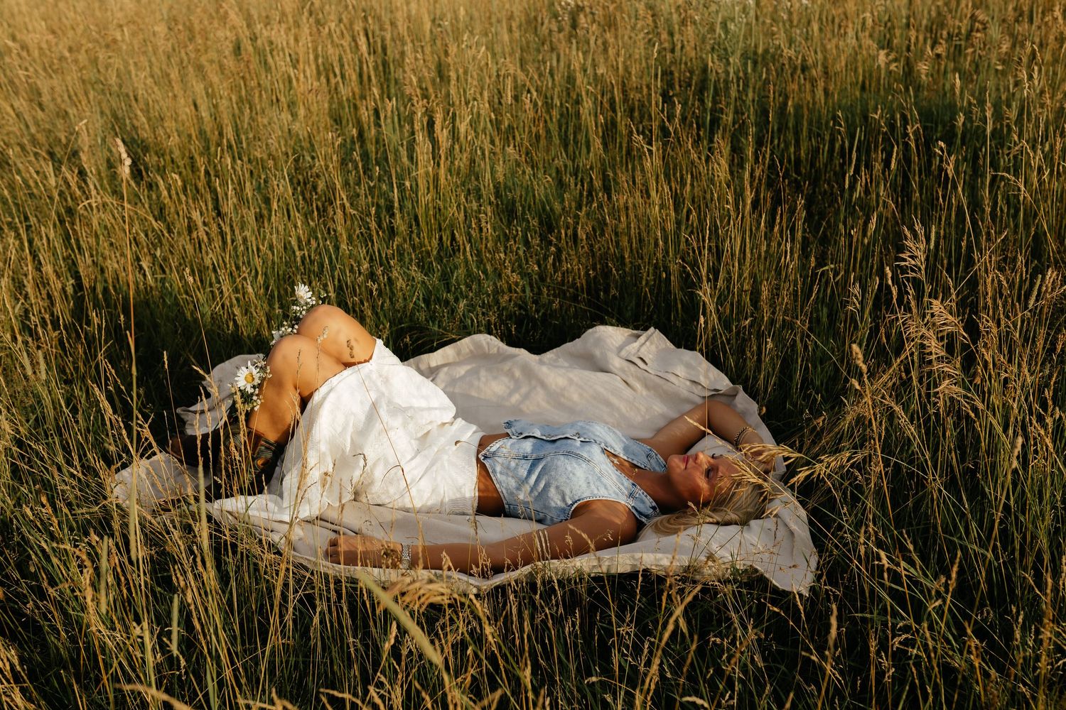 Blonde woman lying on white blanket in tall golden grass field in sunlight.