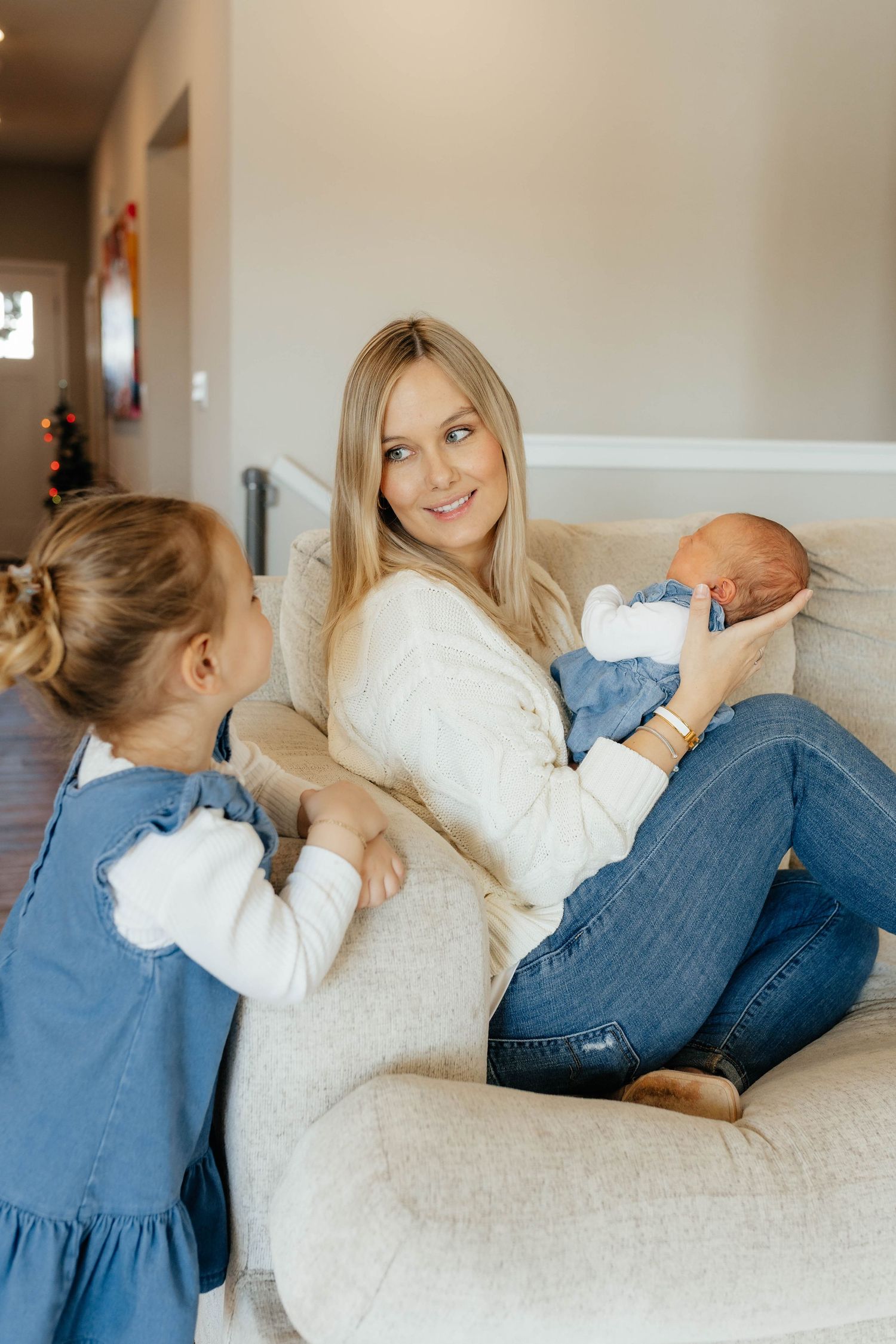 A blonde mother in a white sweater holds her newborn on a chair while her young daughter in denim stands beside her.