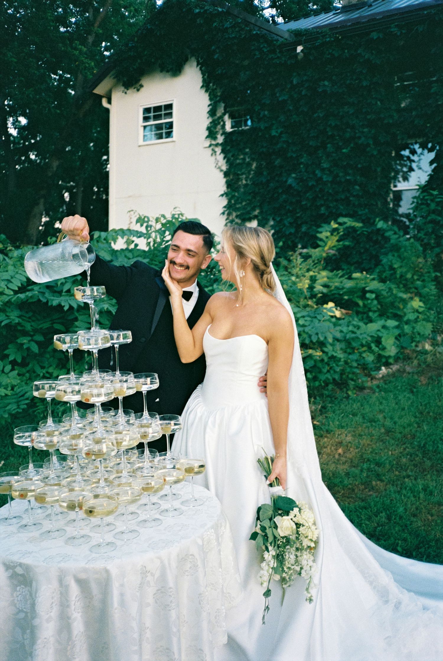 Bride and groom pouring champagne tower at elegant outdoor wedding reception near white building.