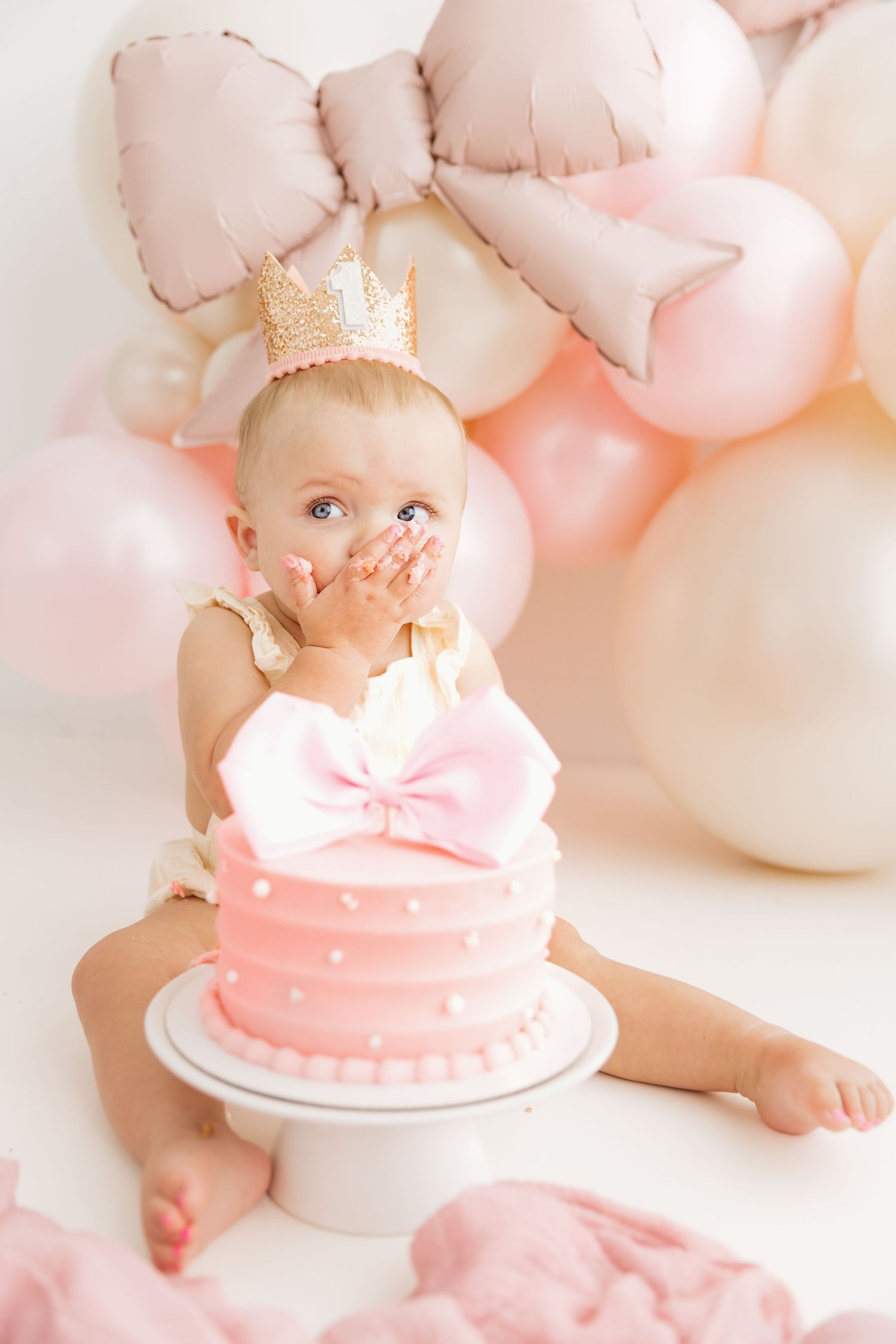 Baby girl in gold crown covers mouth with both hands while sitting beside pink birthday cake.