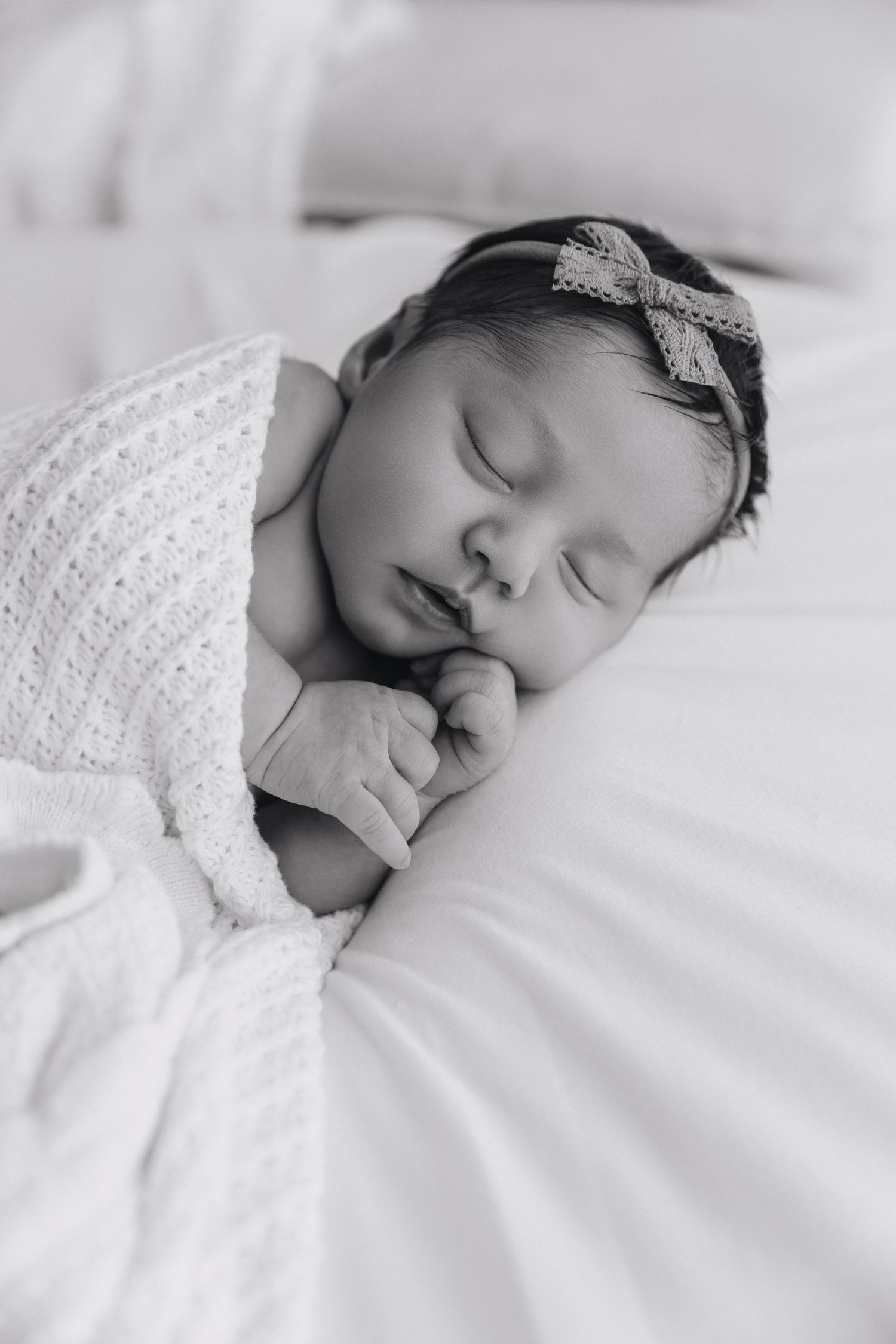Black and white portrait of sleeping newborn baby girl wrapped in white swaddle with lace headband.