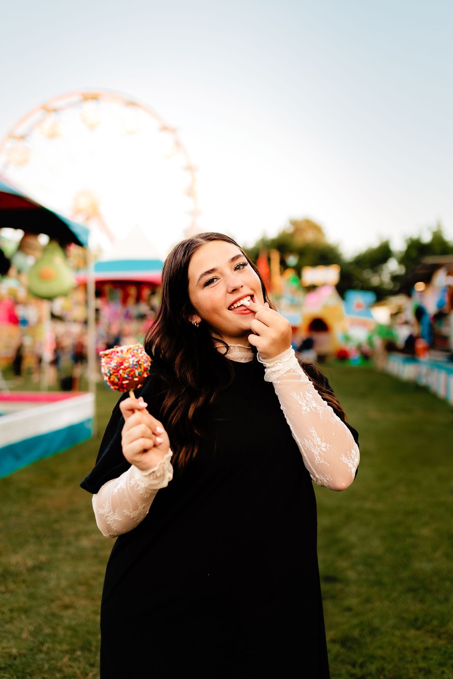 Young woman in black dress smiling while eating a chocolate cake pop at a sunny fairground.