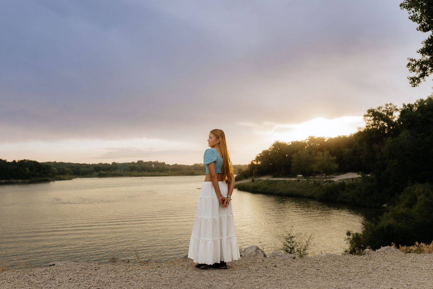 Blonde teen in blue crop top and white maxi skirt stands by lake at golden sunset.