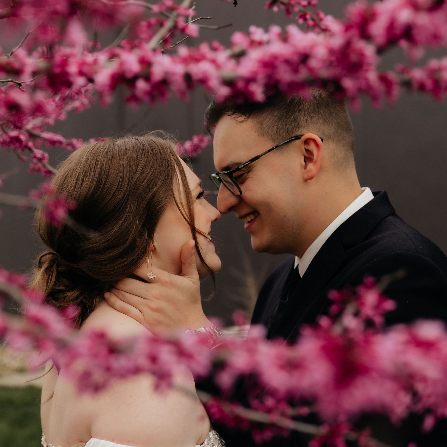 Romantic close-up of bride and groom nearly kissing framed by vibrant pink cherry blossom branches in spring.