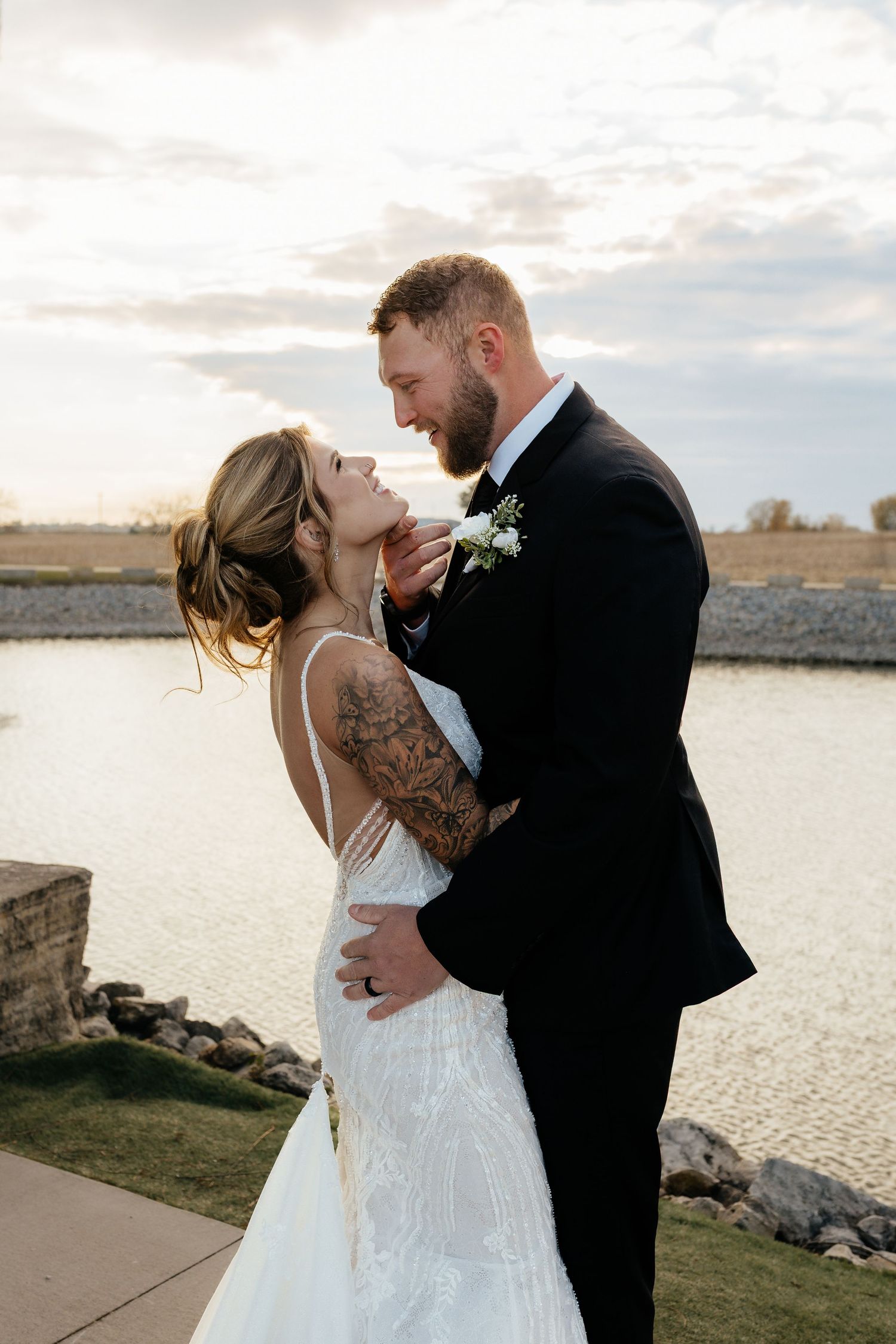 Bride and groom share a romantic moment at sunset near a reflective pond, bride has tattooed arm.