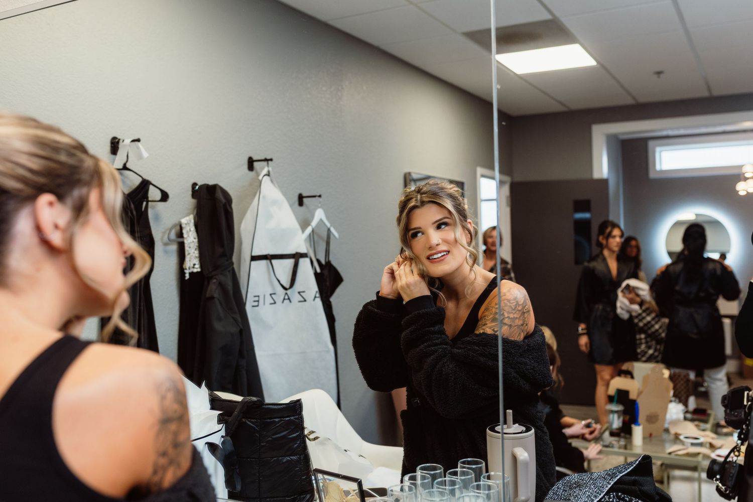 Tattooed bride smiling at her reflection in a salon mirror during wedding day preparations.