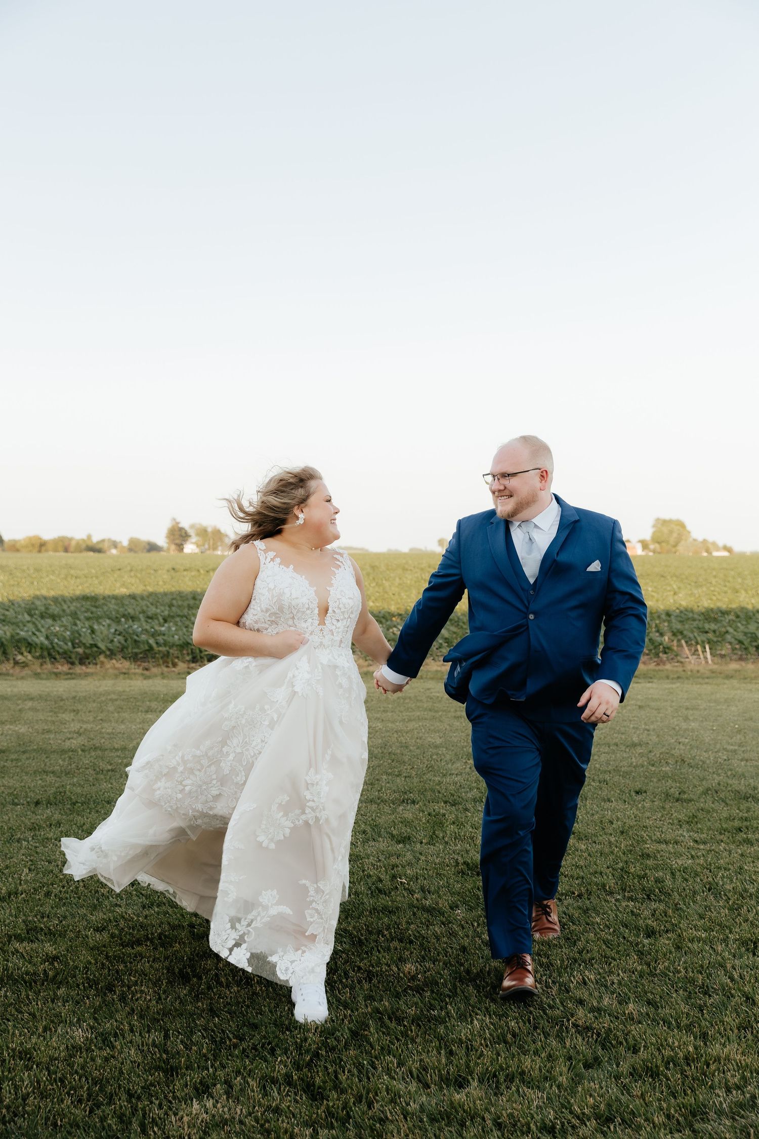 Bride and groom holding hands running through green field, bride in lace gown, groom in navy suit, golden hour.