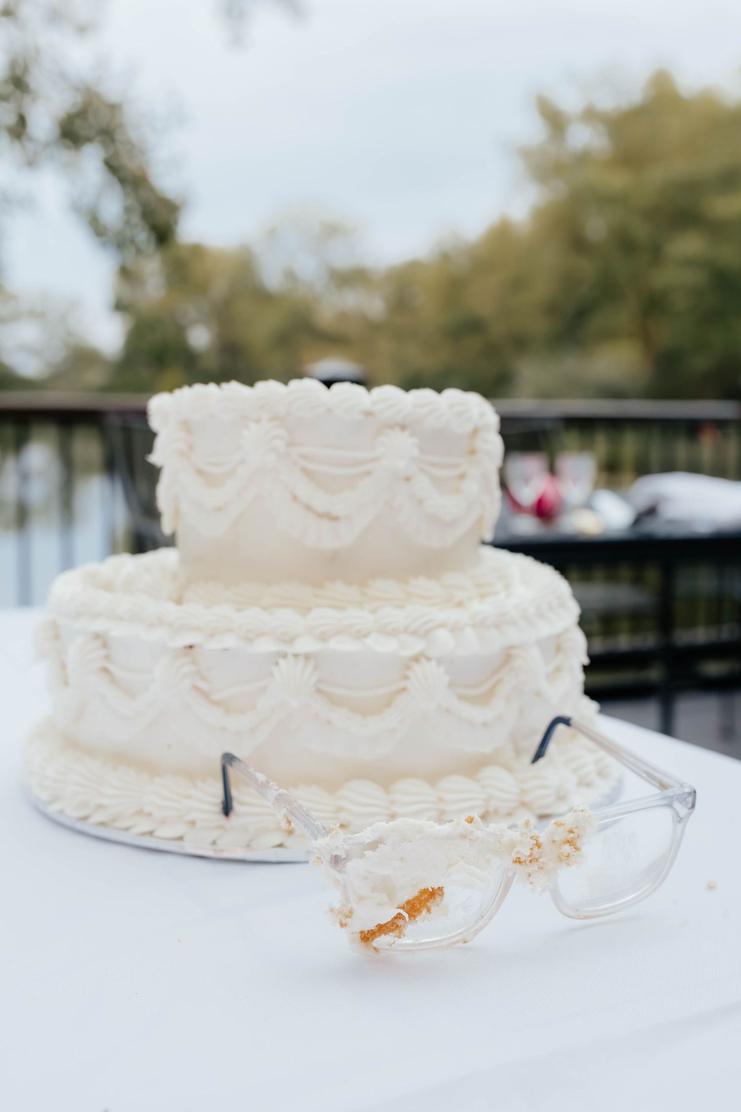 Elegant two-tier white wedding cake with frosting details and glasses resting in front outdoors.