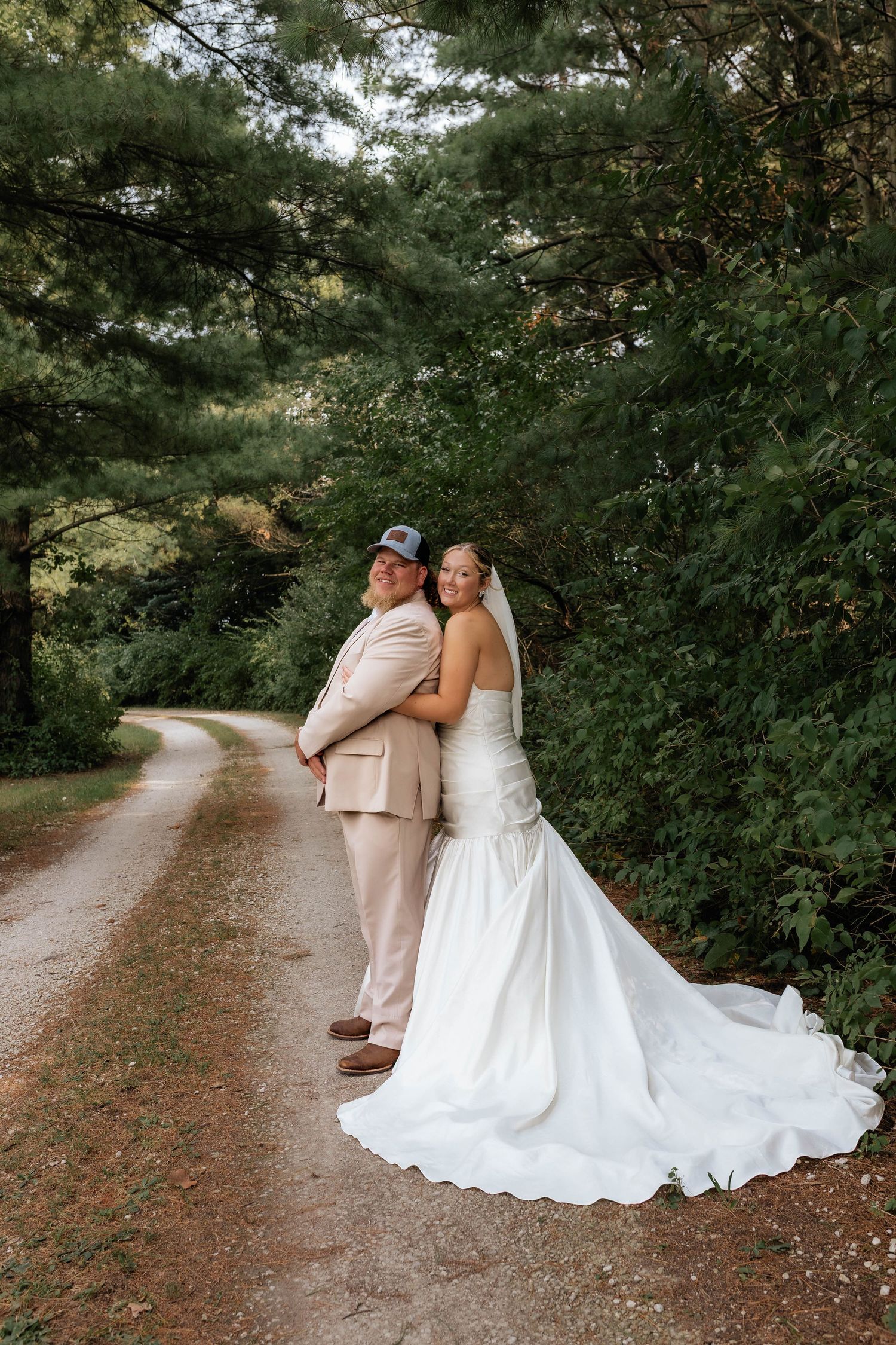 Bride and groom stand back-to-back smiling on a winding wooded path, bride in white gown with train.