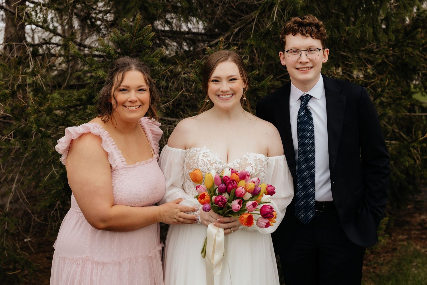 Bride holding colorful tulip bouquet poses with two guests in pink and blue attire.
