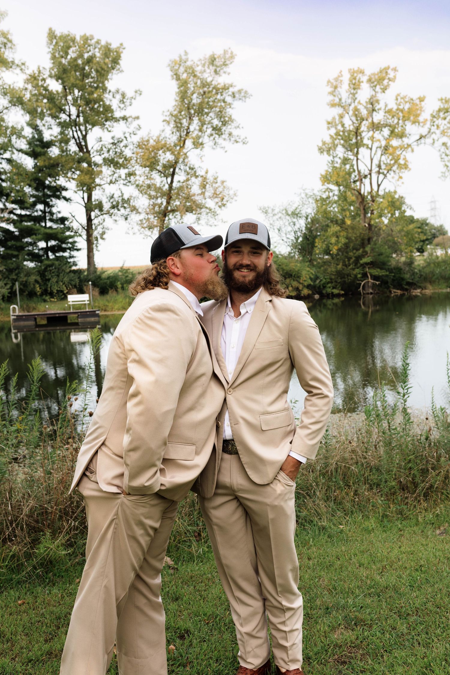 Two groomsmen in tan suits and trucker hats share a playful moment by a scenic pond backdrop.