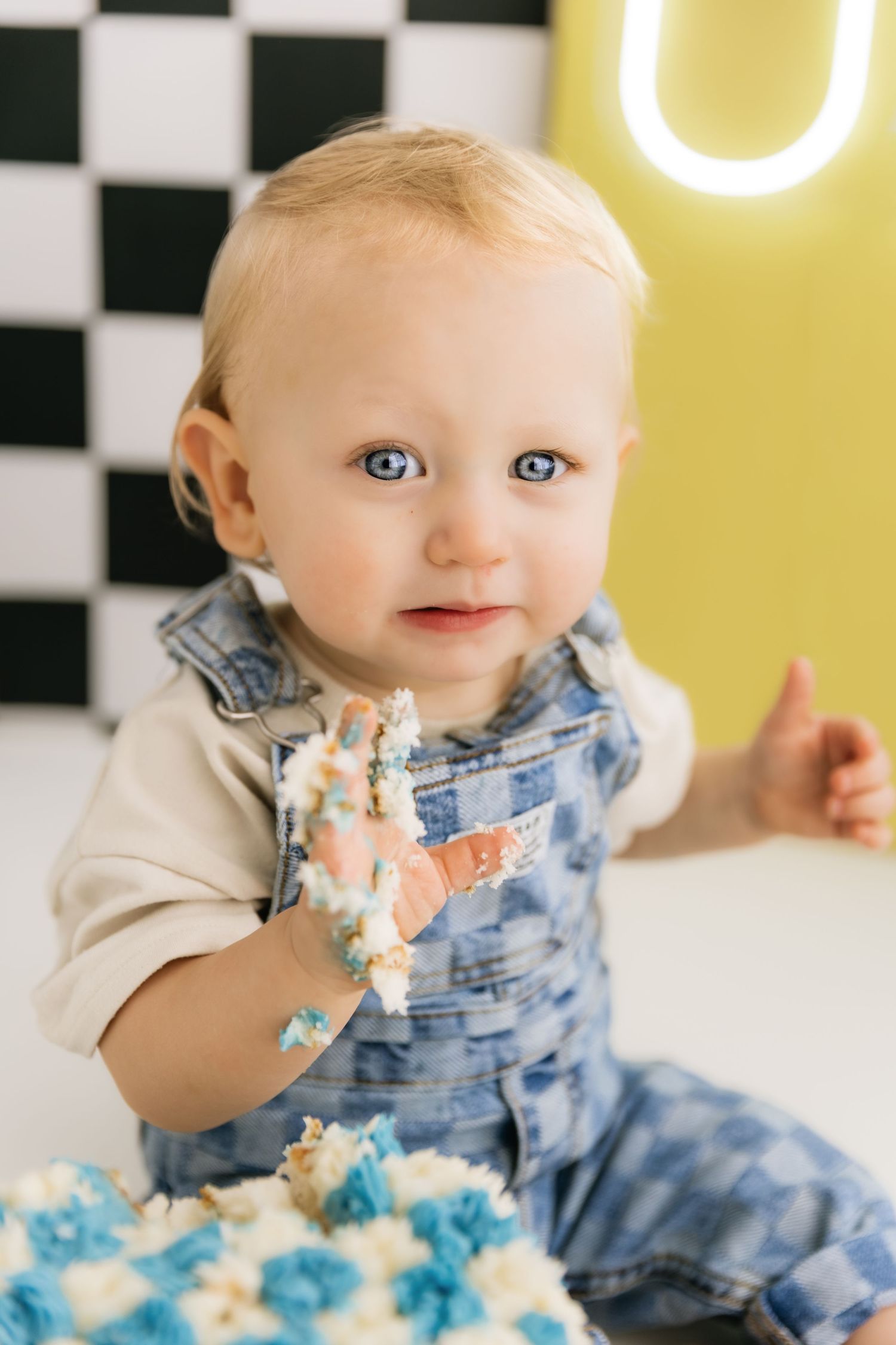 Blue-eyed baby boy in denim overalls with cake frosting on hand at first birthday cake smash celebration.
